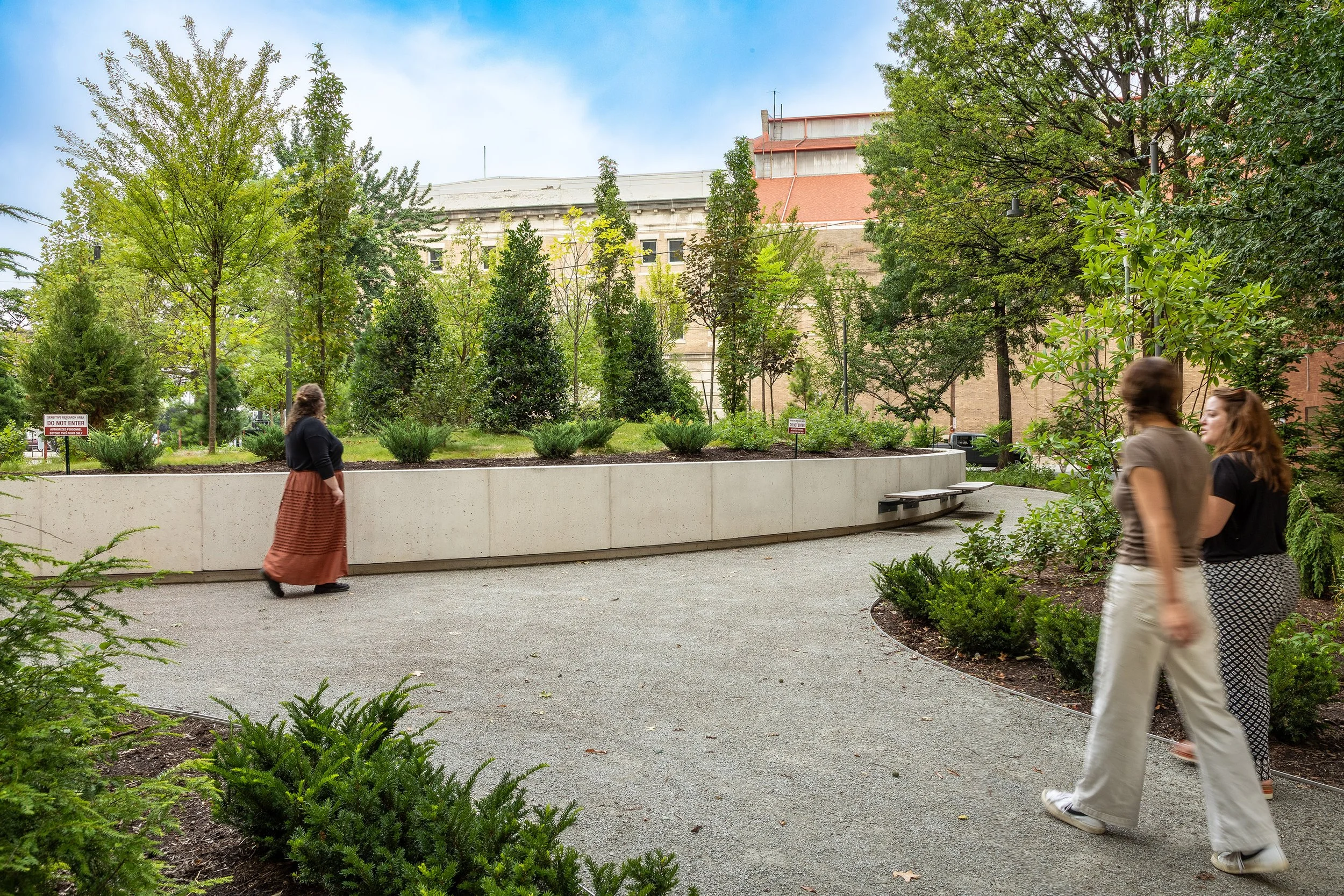 Three people walk along a curved gravel path in an urban landscaped garden with green trees and shrubs, bordered by a low concrete wall. A large building is visible in the background under a partly cloudy sky.