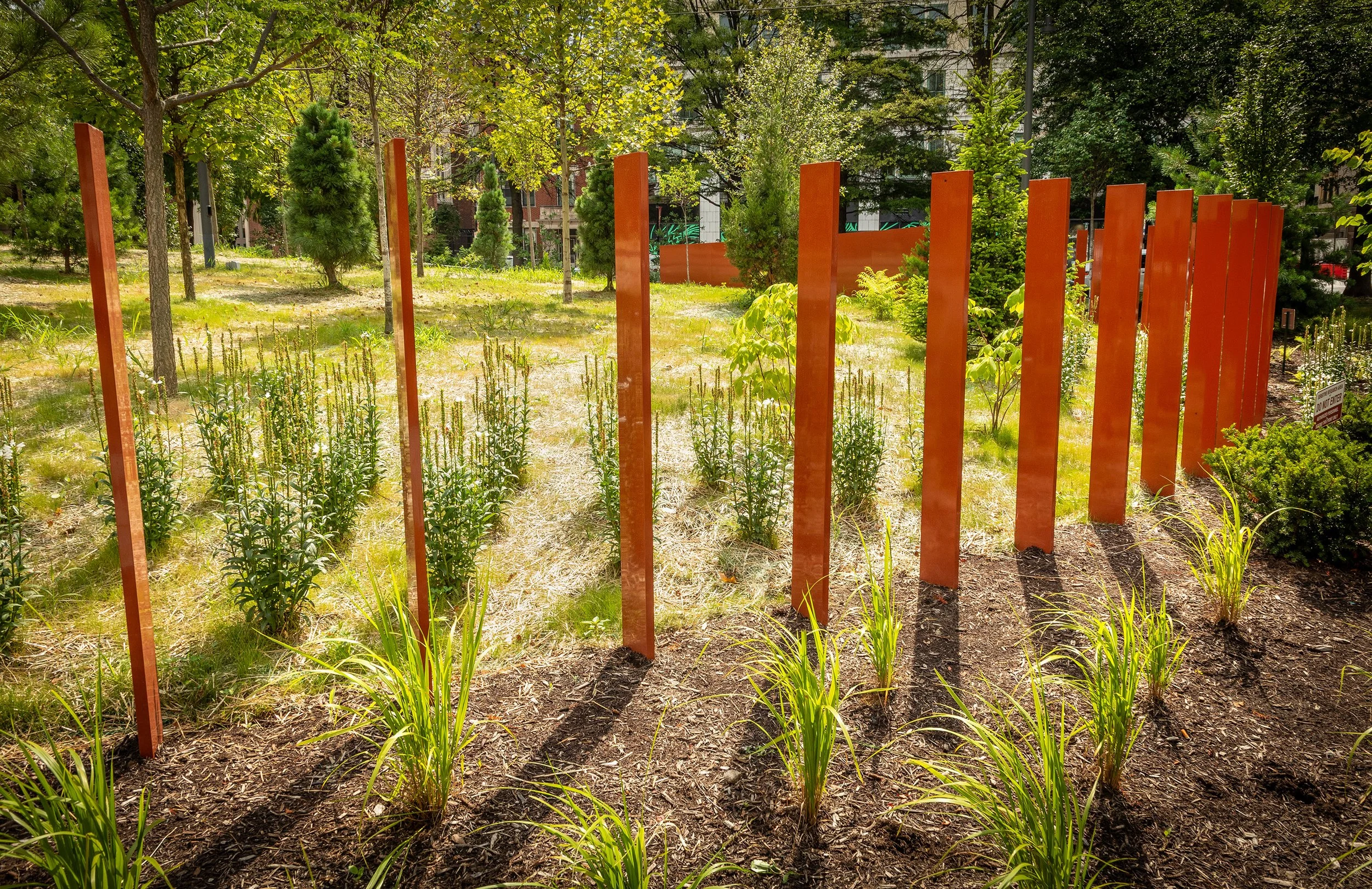 Rows of tall, orange metal beams stand upright among plants and grass in a sunlit park, showcasing striking urban design and casting long shadows on the ground. Trees and greenery fill the background.