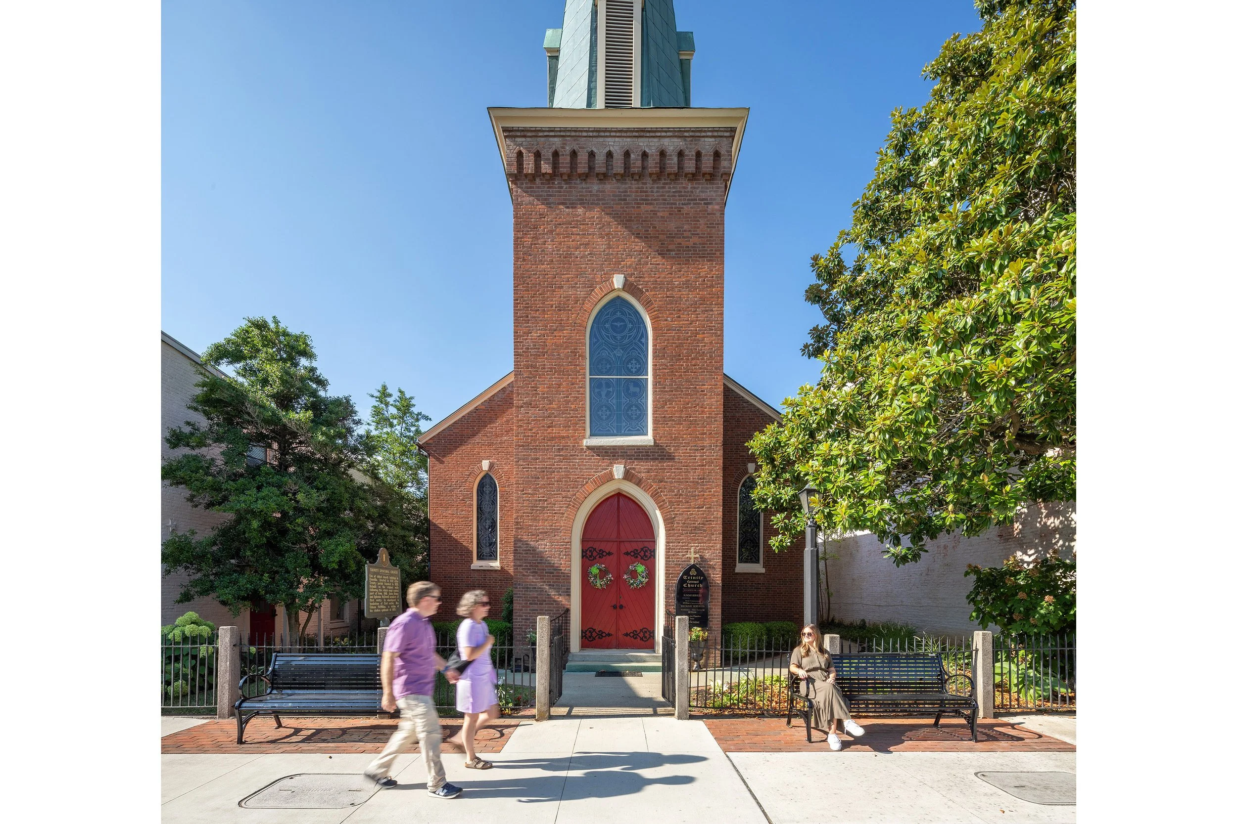A brick church with a steeple, red doors, and arched windows stands along the historic main street as people pass by and one sits on a bench outside.