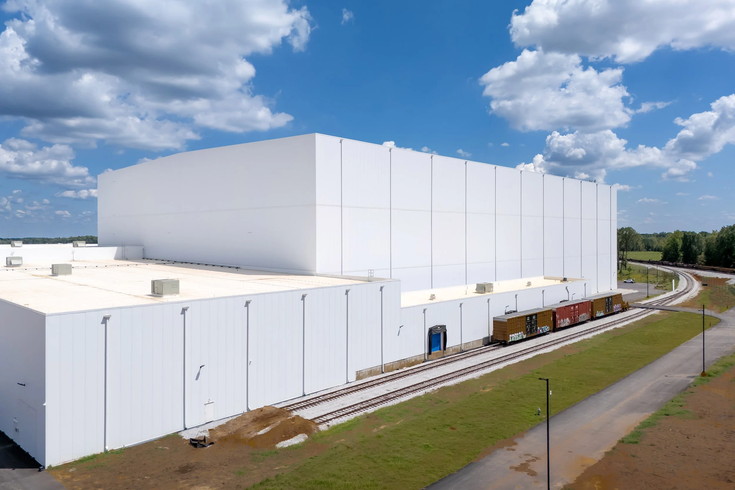 Large white industrial warehouse with refrigeration, loading dock, and train tracks, set under a blue Georgia sky with scattered clouds.