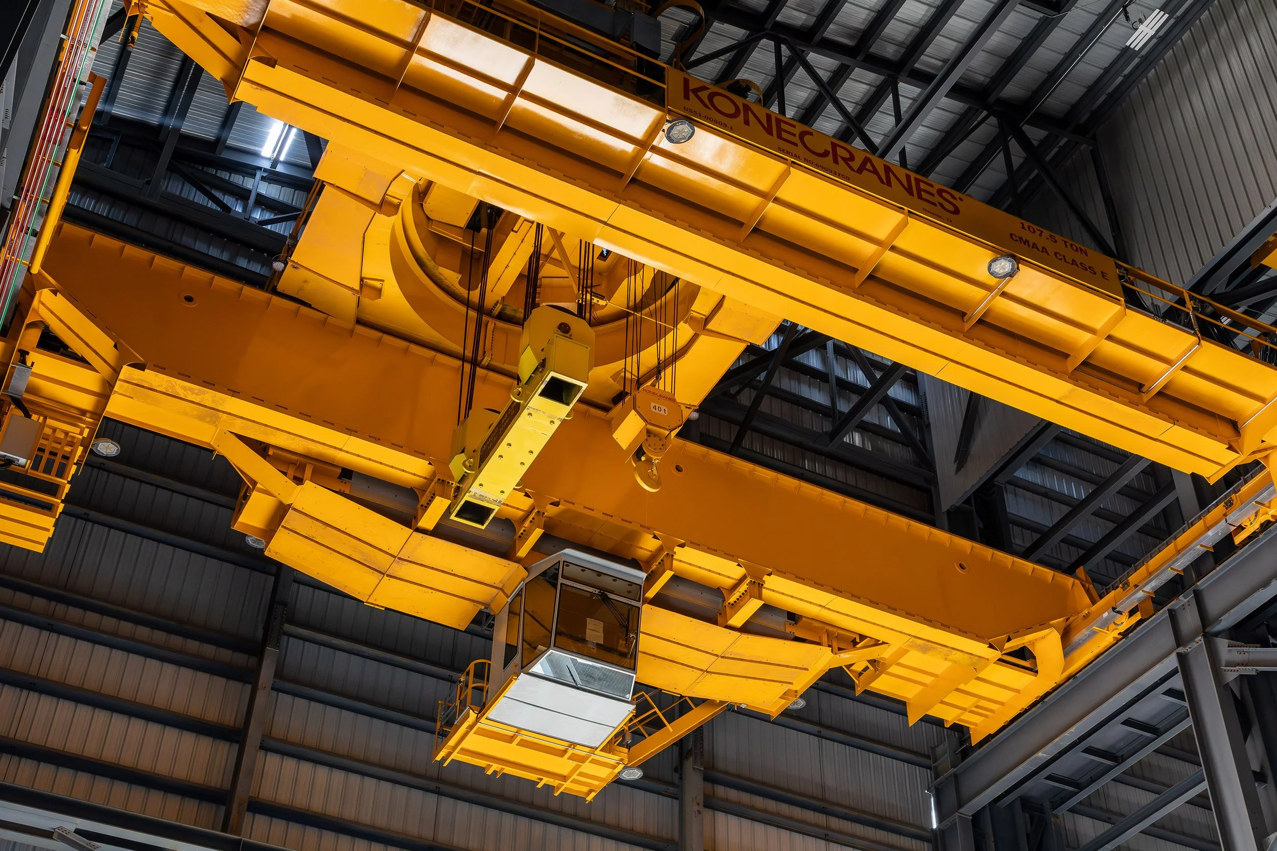 Large yellow overhead crane labeled “Konecranes” inside an industrial warehouse, captured in striking architectural photography, with a control cabin below and surrounded by steel roof structure and metal beams.