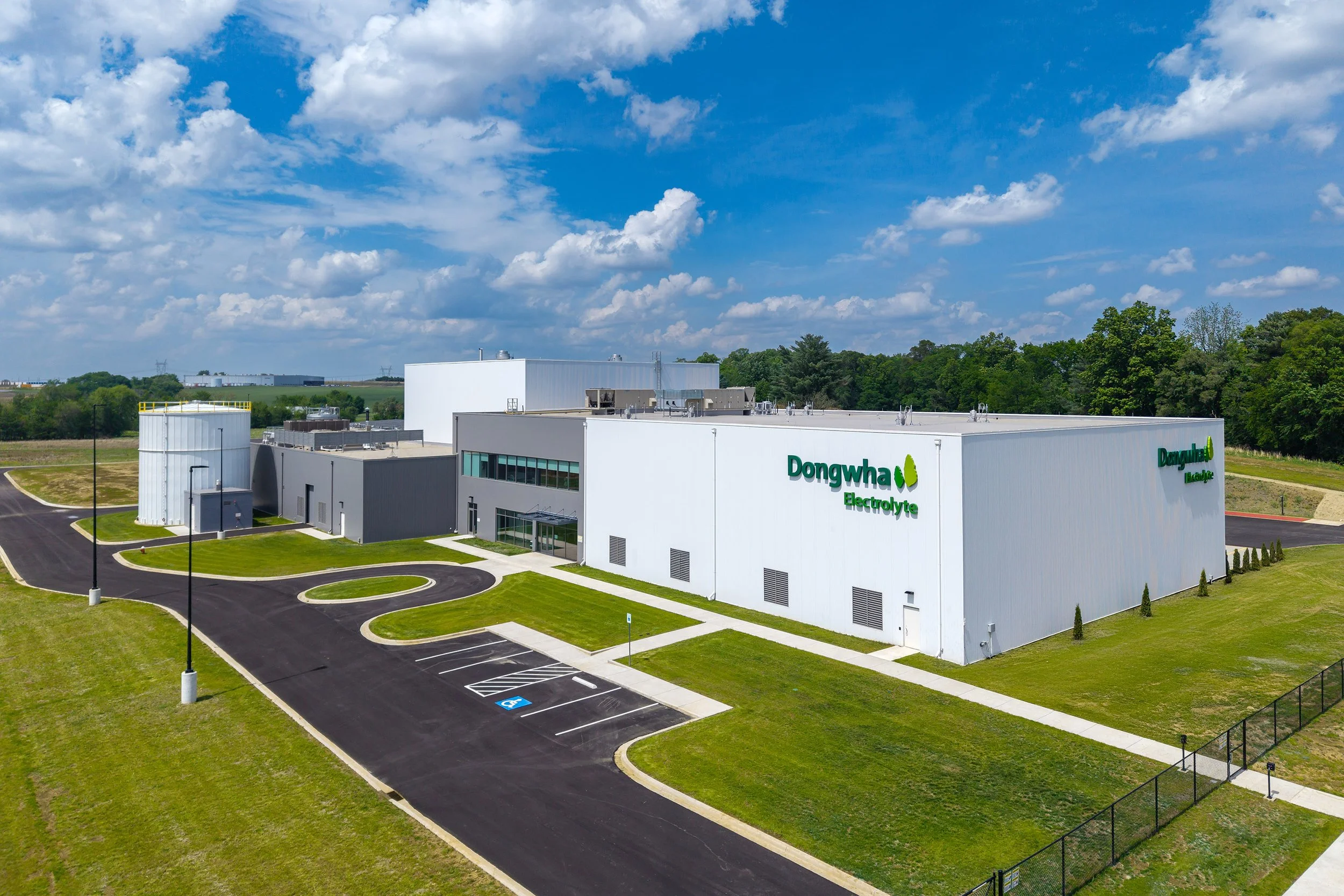A modern white industrial building with Dongwha Electrolyte signage stands amid green grass and trees under a clear blue sky, with a curved driveway and parking spaces in front—perfect example of exceptional architectural photography.
