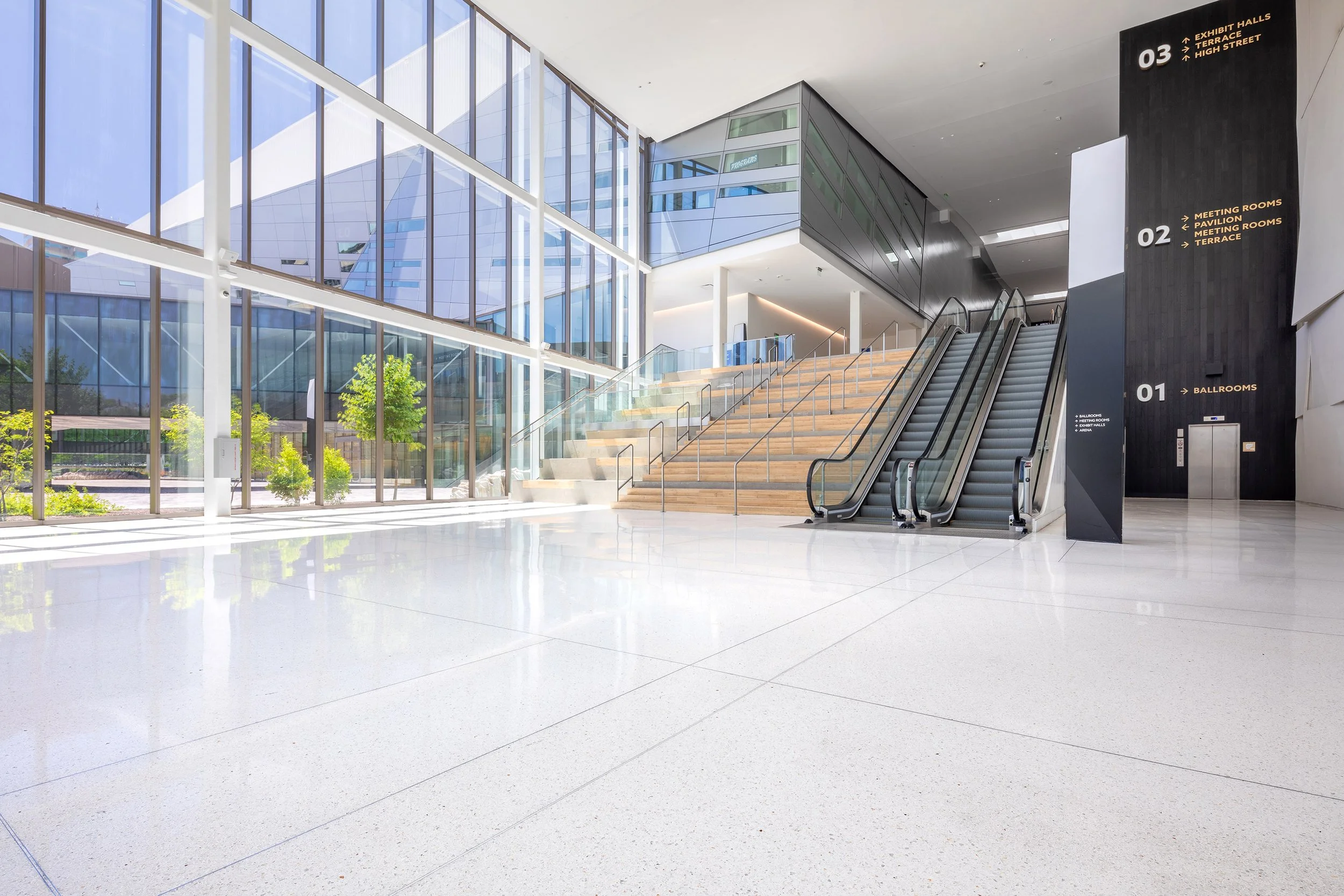 A modern, spacious building lobby featuring terrazzo floors, large windows, an escalator, and directory signage on a black pillar.