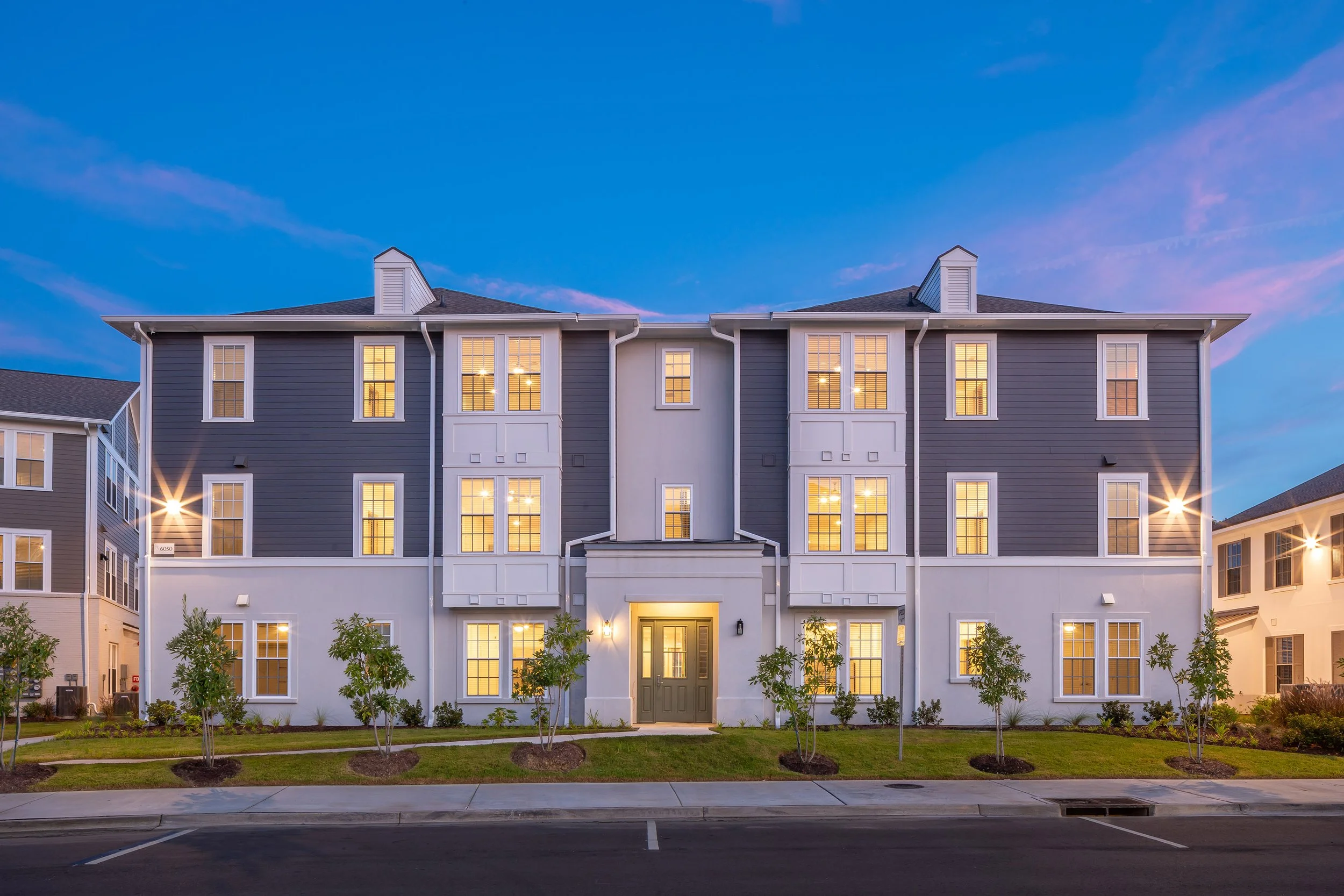 A modern three-story apartment building with gray and white exterior, lit windows, and landscaped shrubs in front, photographed at dusk with a clear blue sky and parking spaces in the foreground.