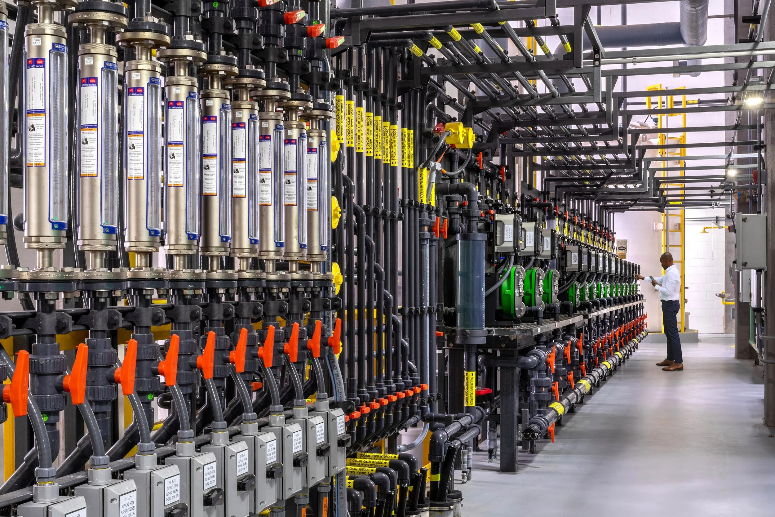 A worker inspecting a complex industrial piping system with various gauges, valves, and pipes in a water treatment facility.