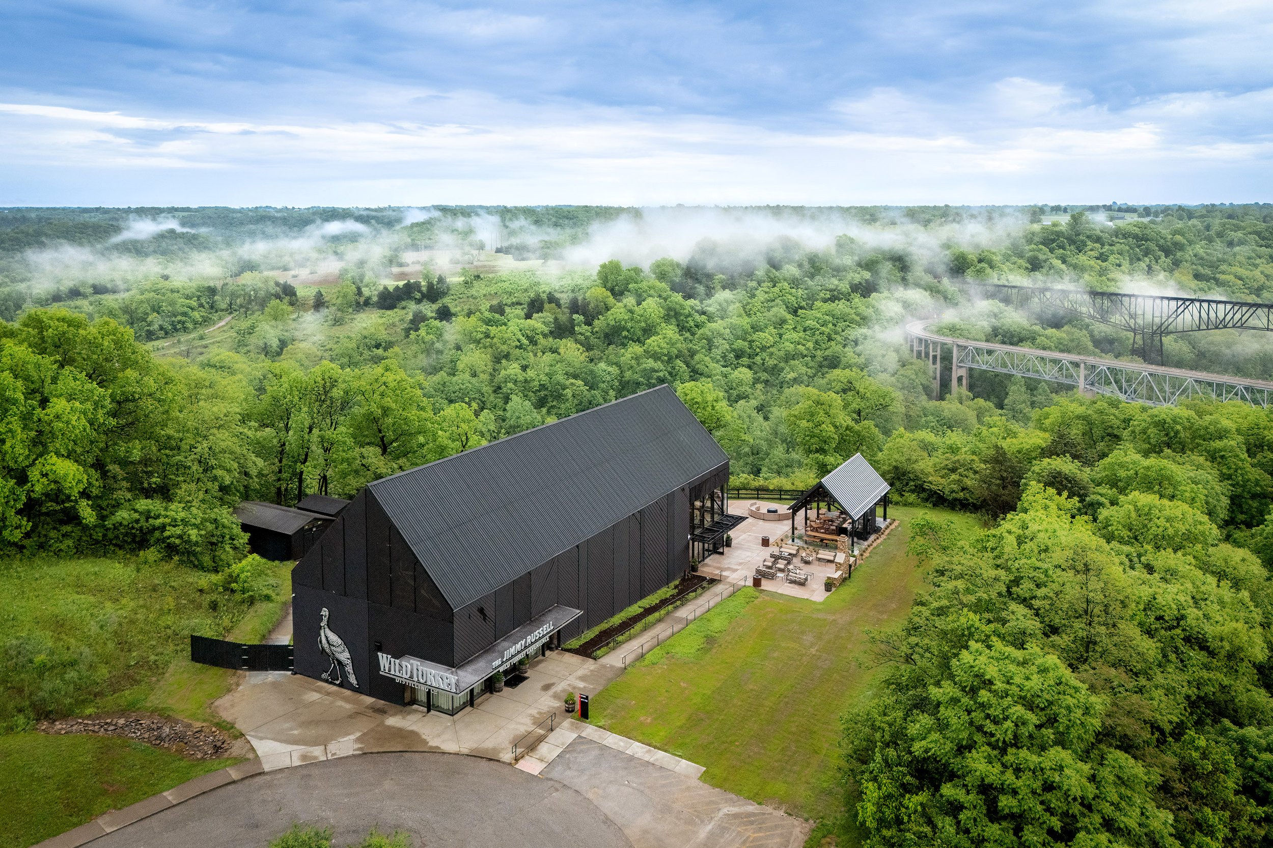 A modern, black industrial distillery labeled “WILD TURKEY” is surrounded by lush green forest and mist, with a bridge visible in the background under a cloudy blue sky.