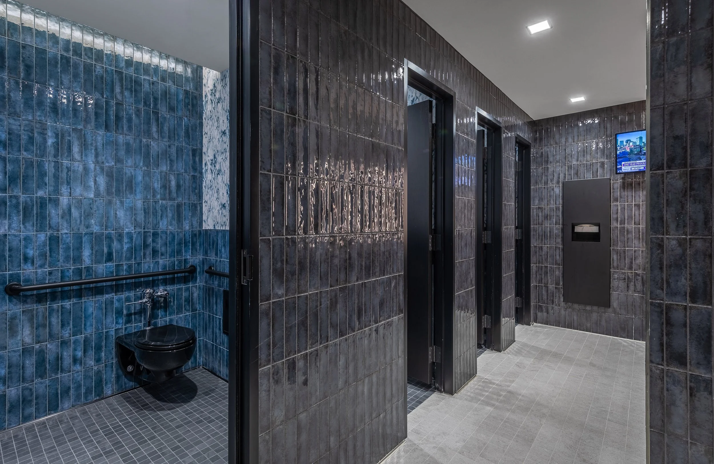 Modern restroom at the arena with dark blue tiled walls, three closed stalls, an open accessible stall with support bars and a black toilet, and bright overhead lighting. A small digital screen is mounted on the wall near the sink area.