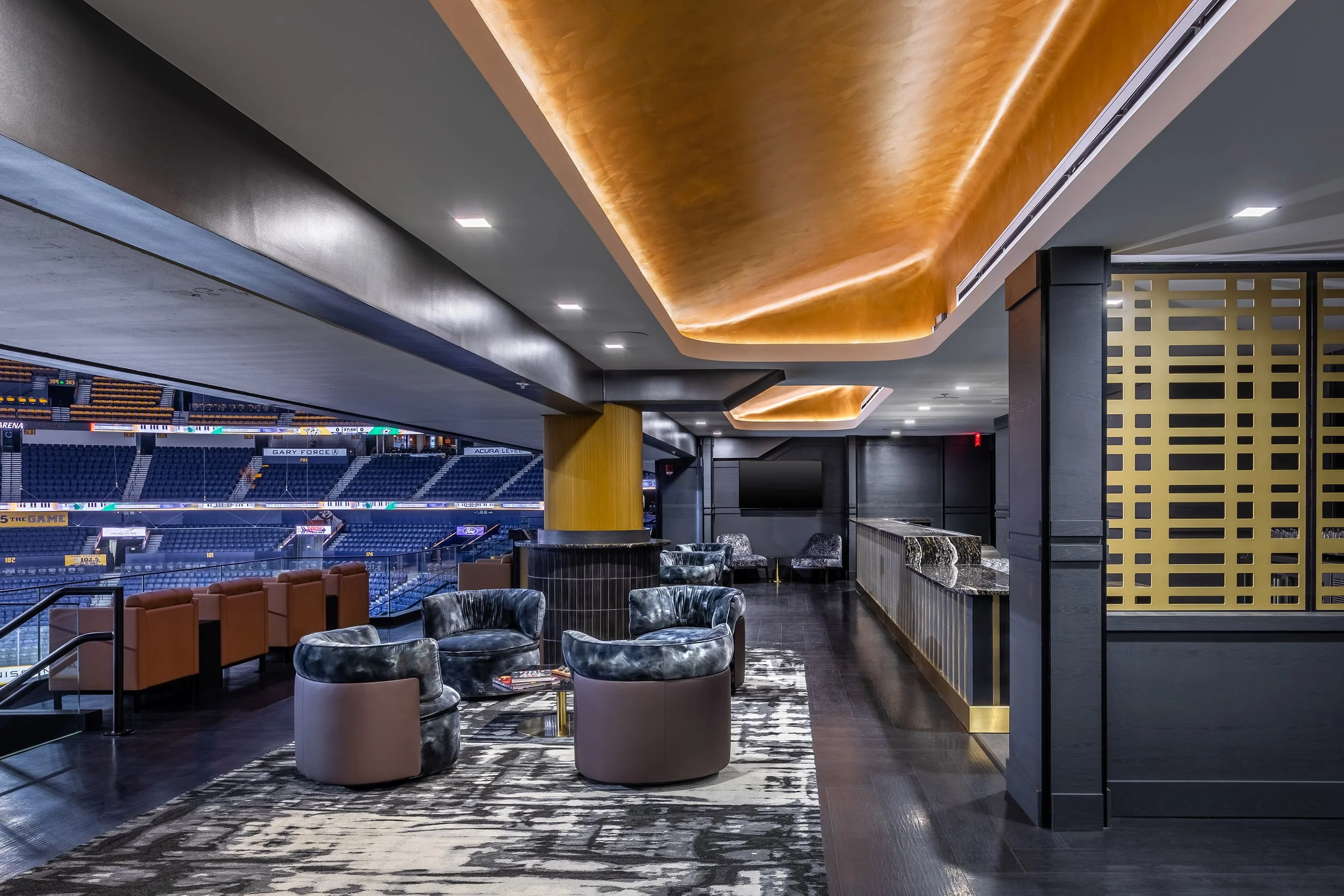 Modern luxury suite at a sports arena featuring stylish seating, a patterned rug, sleek bar area, and expansive views of the arena seats and scoreboard from the large open terrace. Warm lighting and contemporary design details throughout.