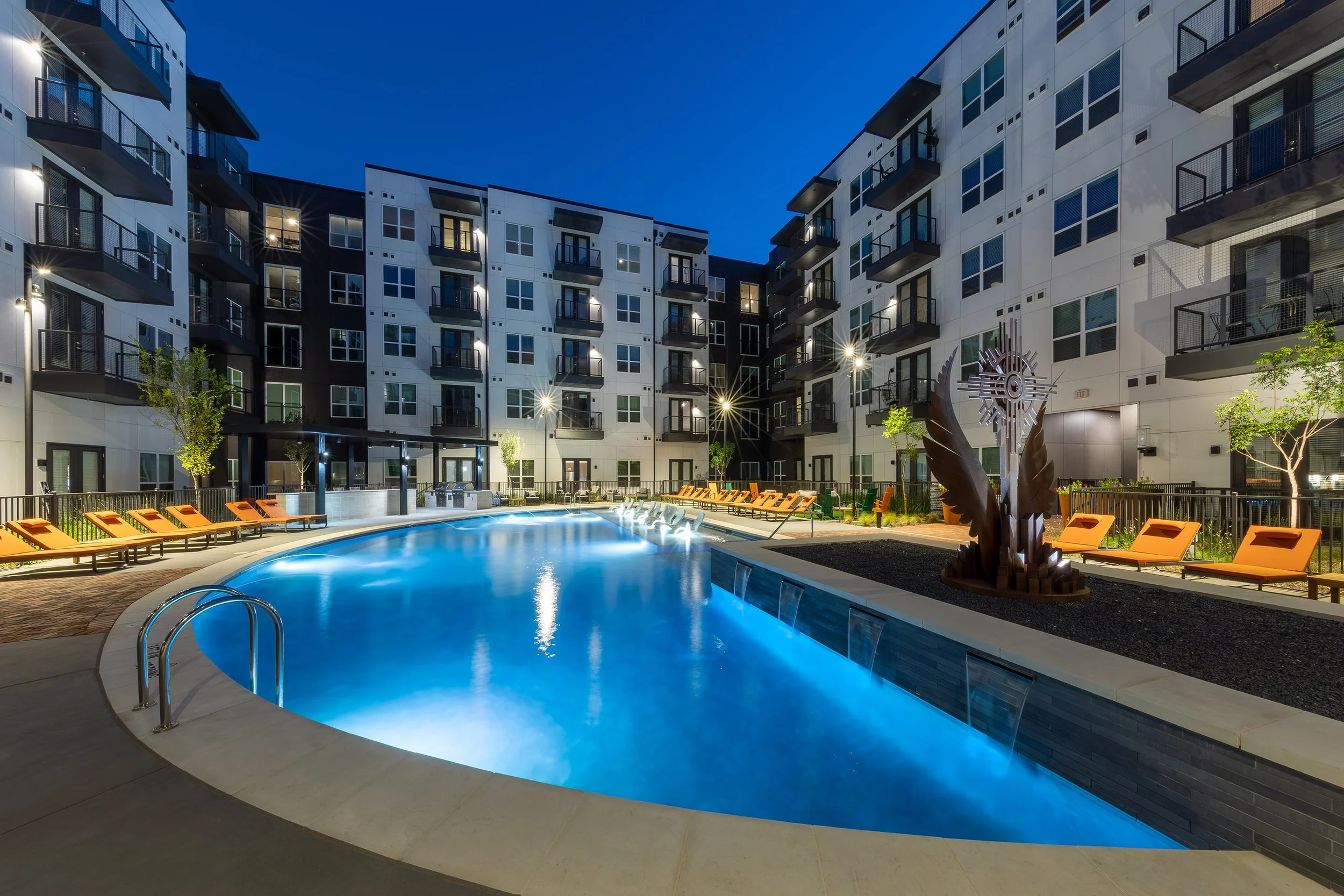 Modern apartment complex at night with a well-lit, curved swimming pool surrounded by orange lounge chairs, trees, and contemporary sculptures; multiple stories of balconies overlook the pool area.