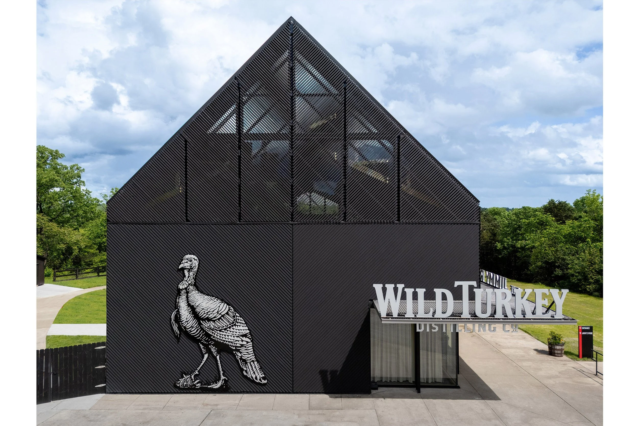 A modern, black distillery building with striking architecture features a large metal wild turkey sculpture and the sign Wild Turkey Distilling Co. at the entrance, surrounded by trees under a partly cloudy sky.