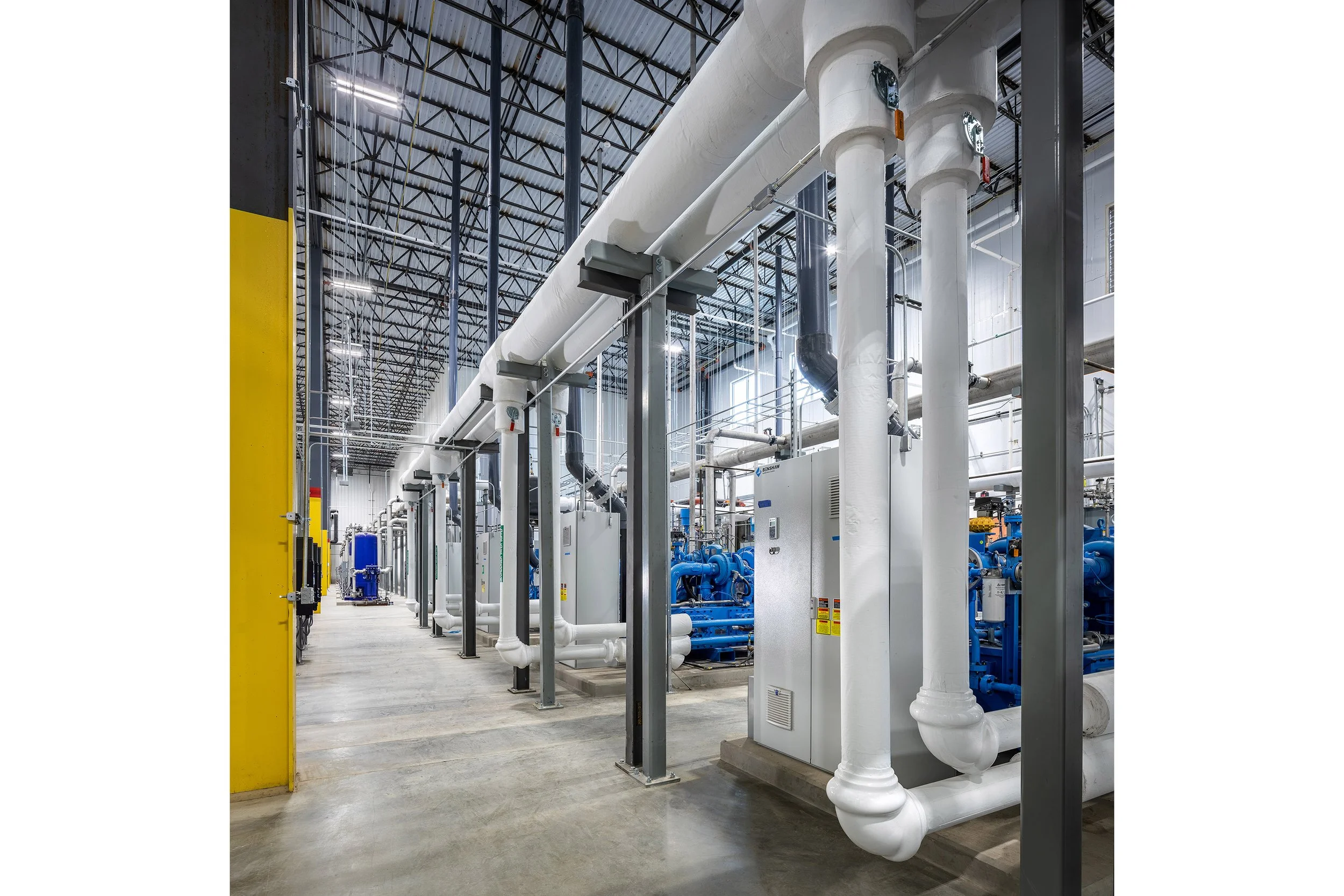 Industrial facility with large white insulated process pipes, metal beams, and machinery arranged in neat rows on a concrete floor under a high, open ceiling with exposed lighting and beams.