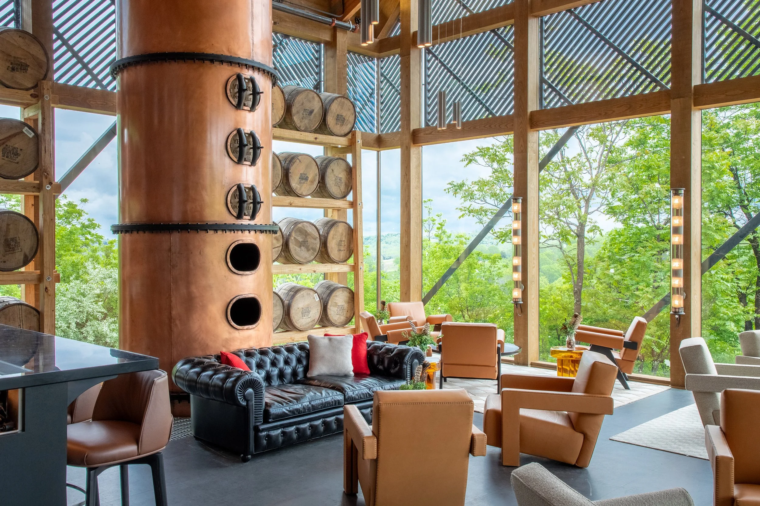 A modern lounge with leather chairs, a black tufted sofa, a tall copper distillery column, stacked bourbon barrels, and large windows overlooking green trees and hills—perfect for relaxing at the Wild Turkey visitor center.