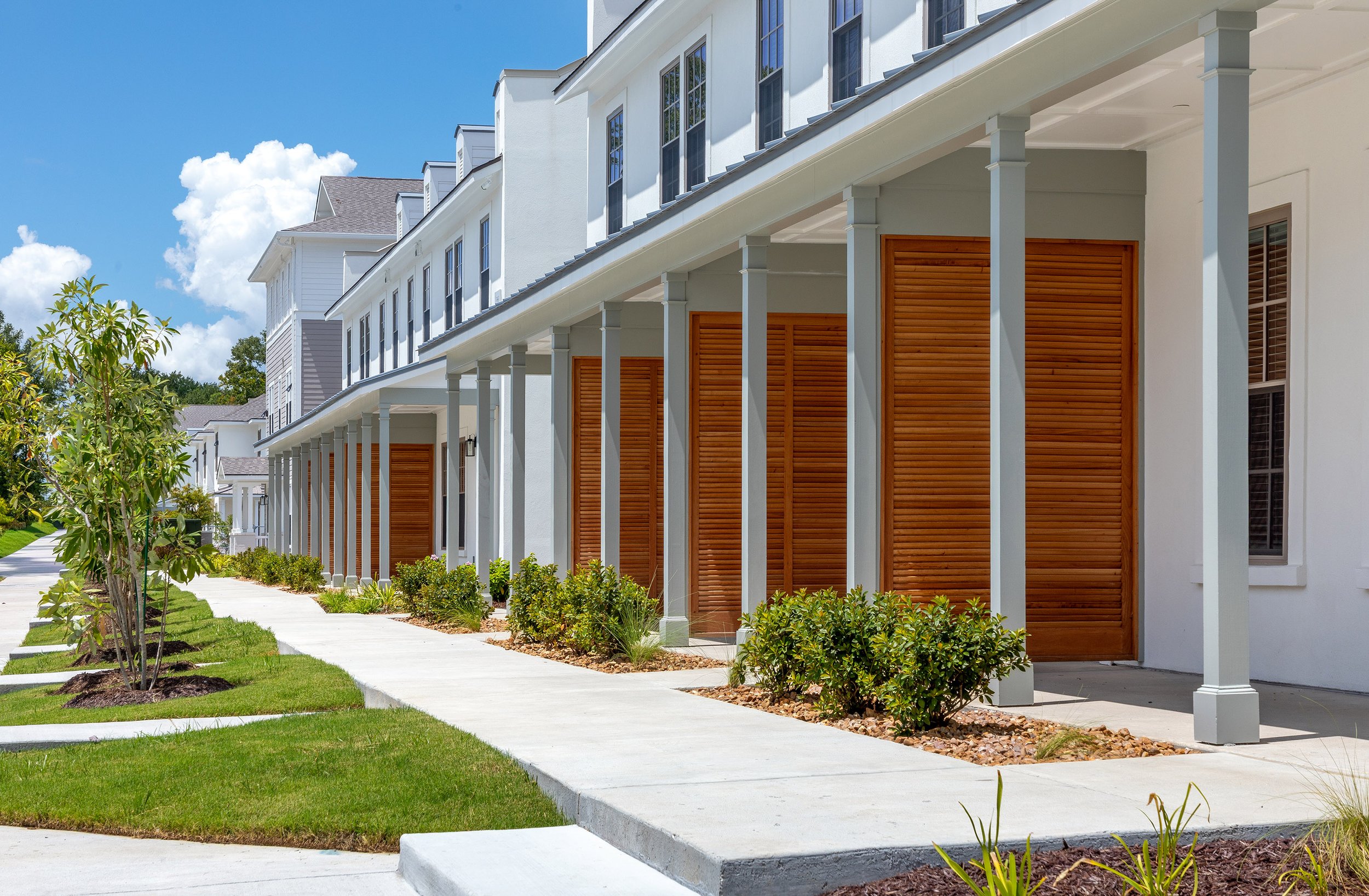 A row of modern white townhouses with tall wooden doors, covered porches supported by columns, and neatly landscaped walkways lined with shrubs and grass under a bright blue sky.