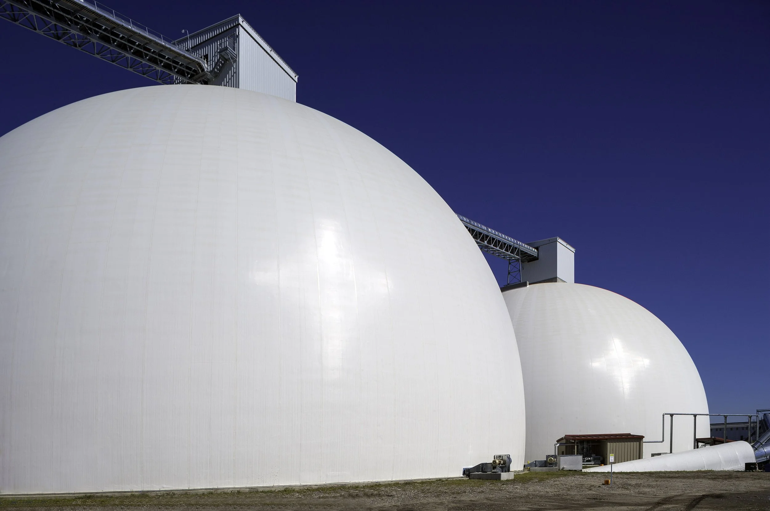Two large white storage domes under a clear blue sky, with conveyor structures and small buildings nearby.