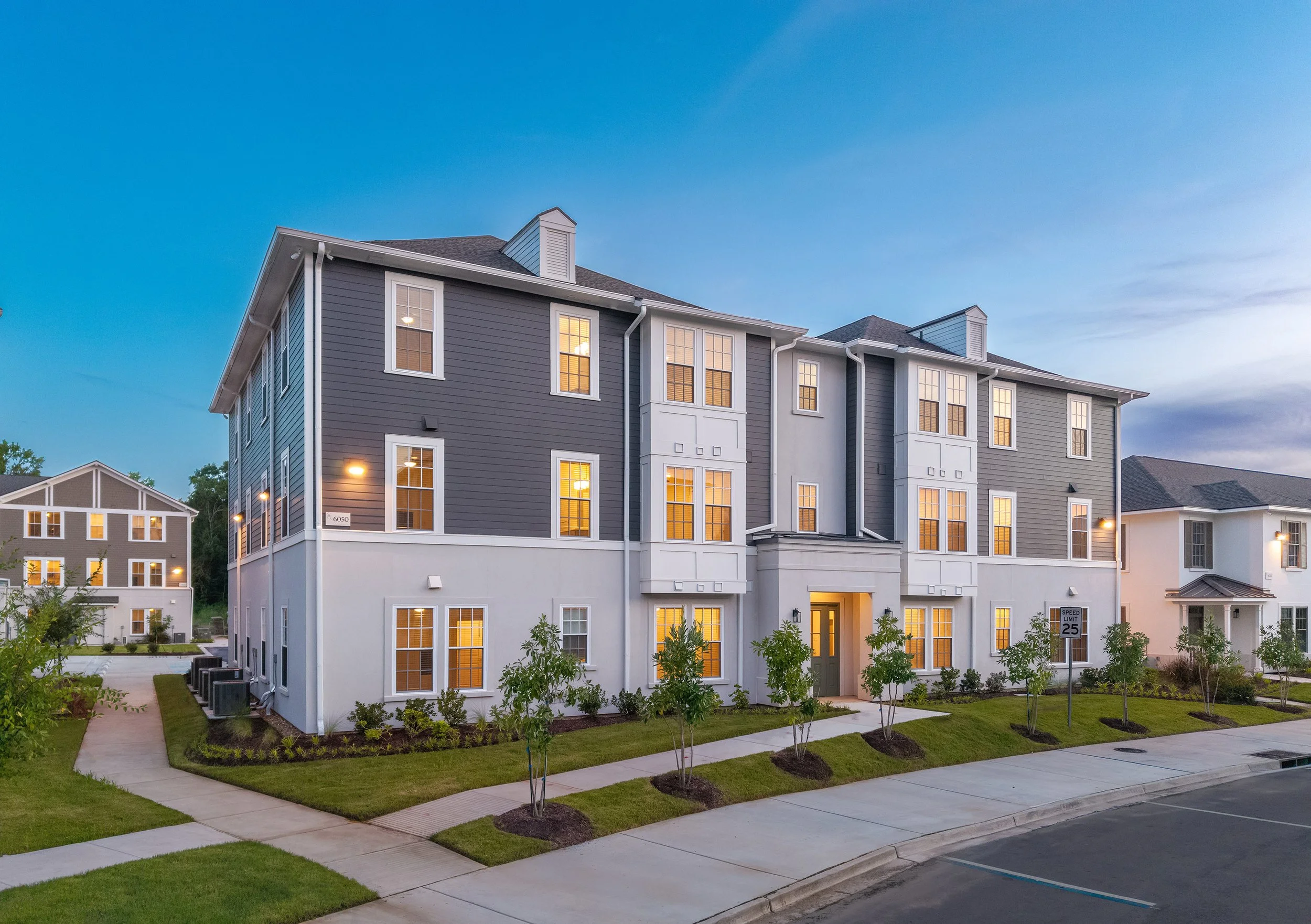 Modern three-story apartment building with gray and white siding, large windows lit from within, landscaped lawns, and a clear evening sky in the background. A sidewalk and street are visible in the foreground.