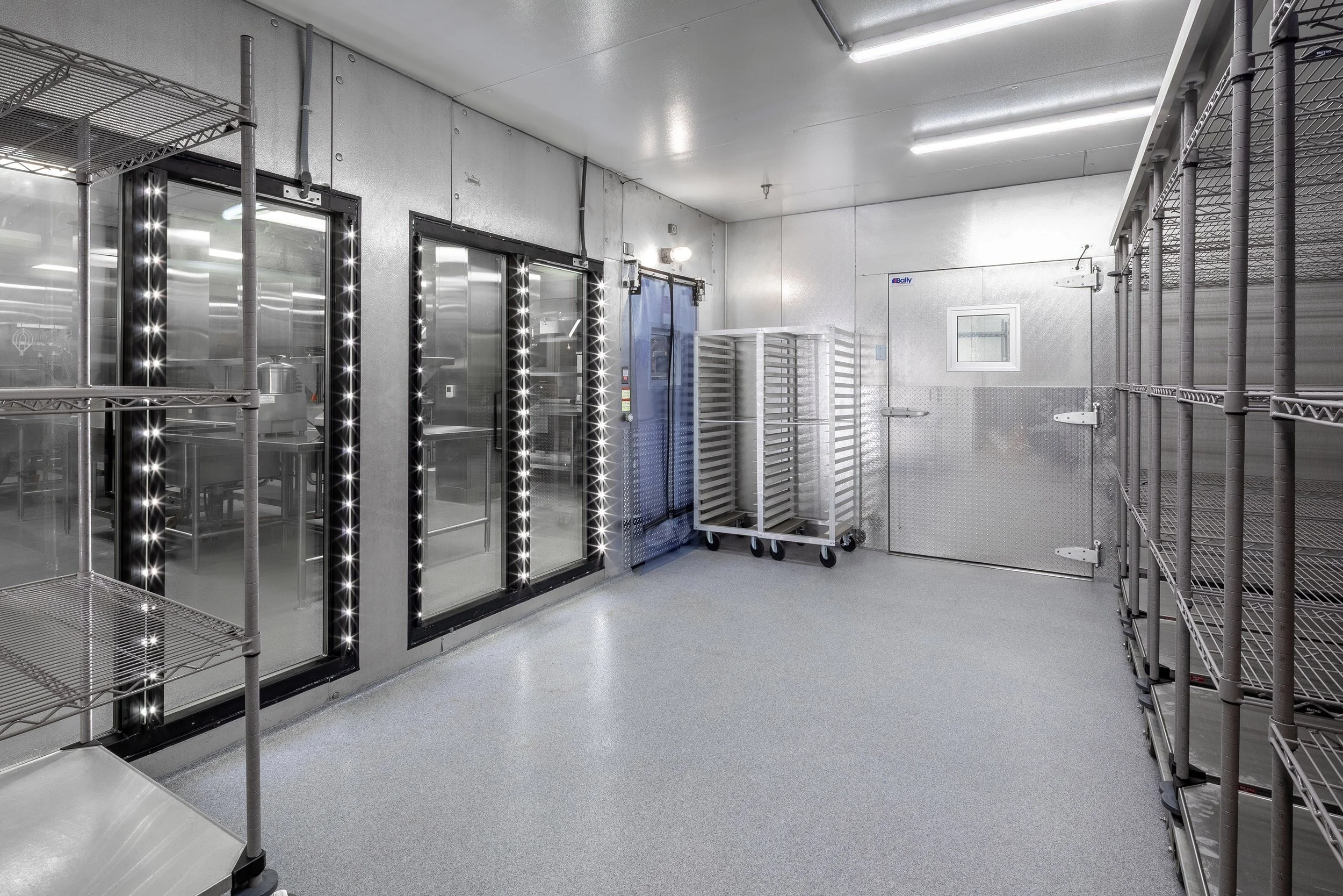 A clean, modern commercial walk-in refrigerator with metal shelves, glass doors, and several empty rolling racks sits on a gray floor under bright ceiling lights—ideal for healthcare interiors demanding hygiene and efficiency.