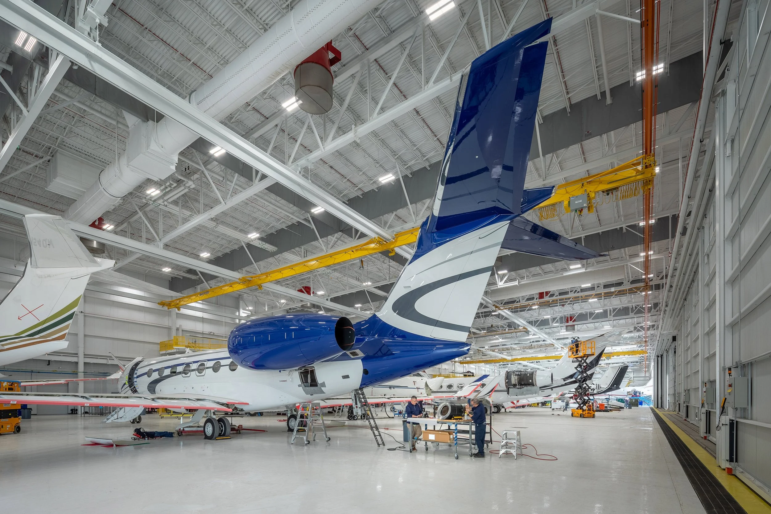 A large, well-lit aircraft hangar in Arizona with several private jets being serviced. The focus is on a blue and white jet, surrounded by maintenance equipment and workers. Architectural details and other aircraft fill the spacious construction.