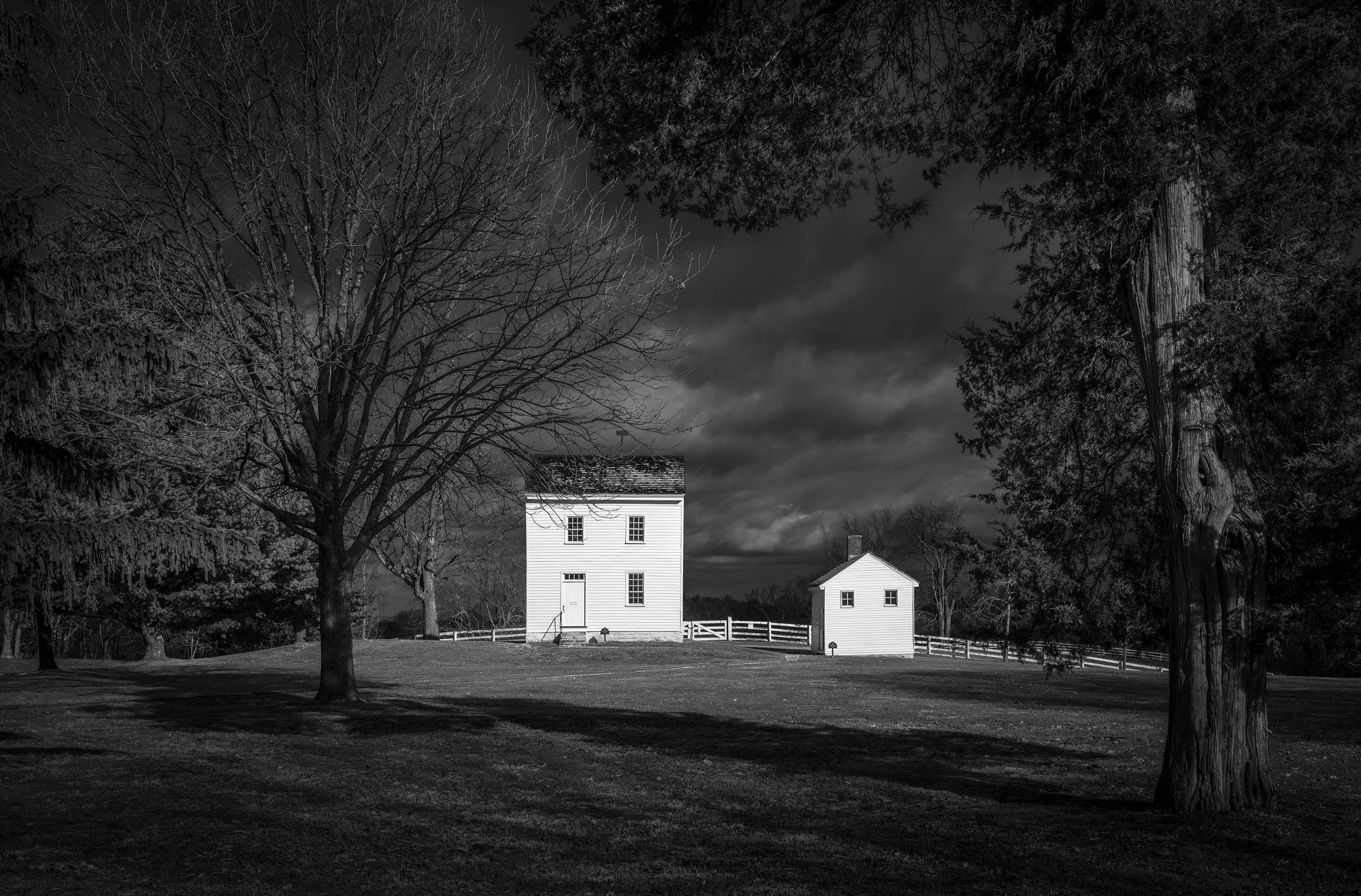 Two white Shaker buildings stand in a grassy field, framed by bare trees under a dramatic winter sky.