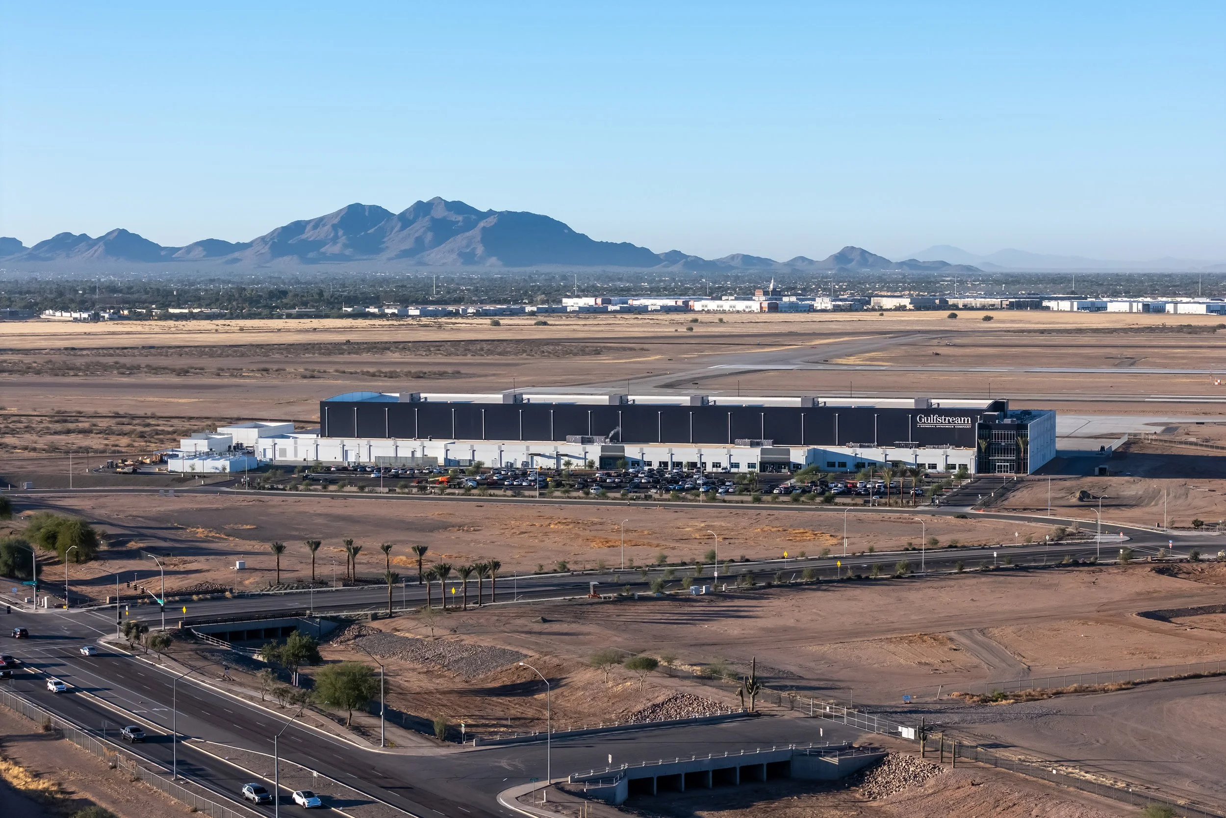 An aerial photography view of a large industrial building in the Arizona desert, with mountains in the background and parking lots and roads surrounding the facility situated near an airport runway.