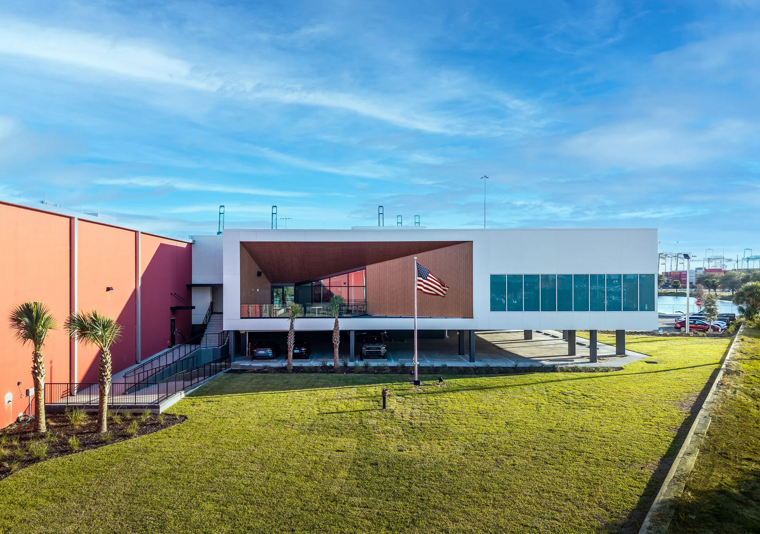 A modern commercial building with a red and white exterior, large windows, and a covered parking area sits behind a lawn with a flagpole displaying the American flag under a blue sky.