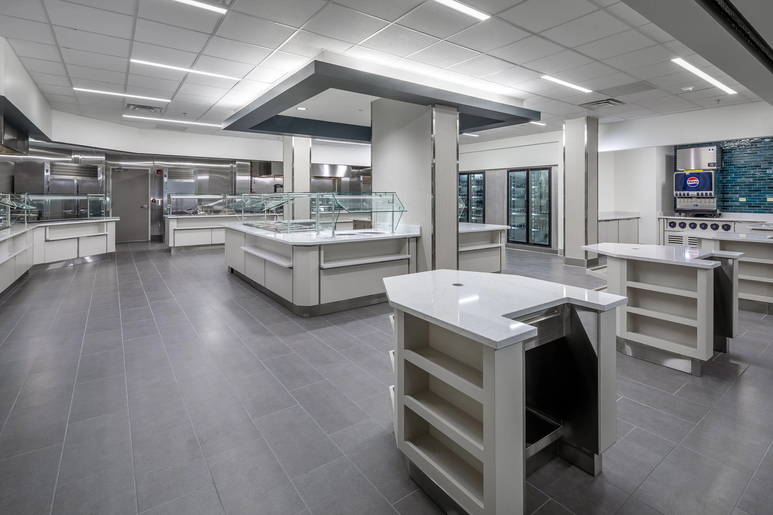 A modern, empty commercial kitchen designed for interiors photography, featuring multiple serving stations, stainless steel appliances, glass food display cases, and gray tile flooring under bright ceiling lights.