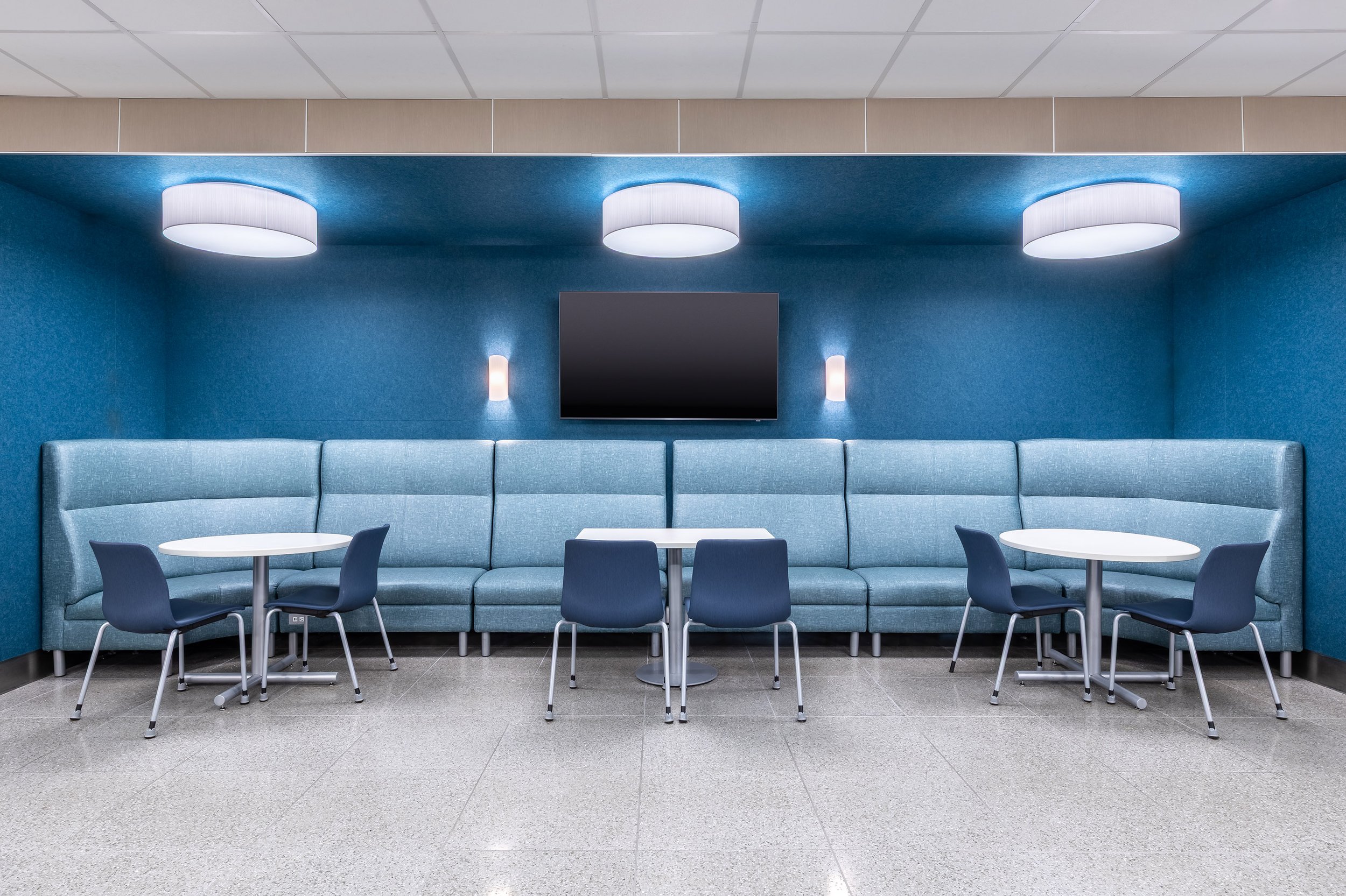 A modern, blue-accented lounge in a healthcare setting with long upholstered bench seating, three round white tables, blue chairs, wall-mounted TV, and circular ceiling lights. The shiny gray tile floor adds to the clean and welcoming ambiance.