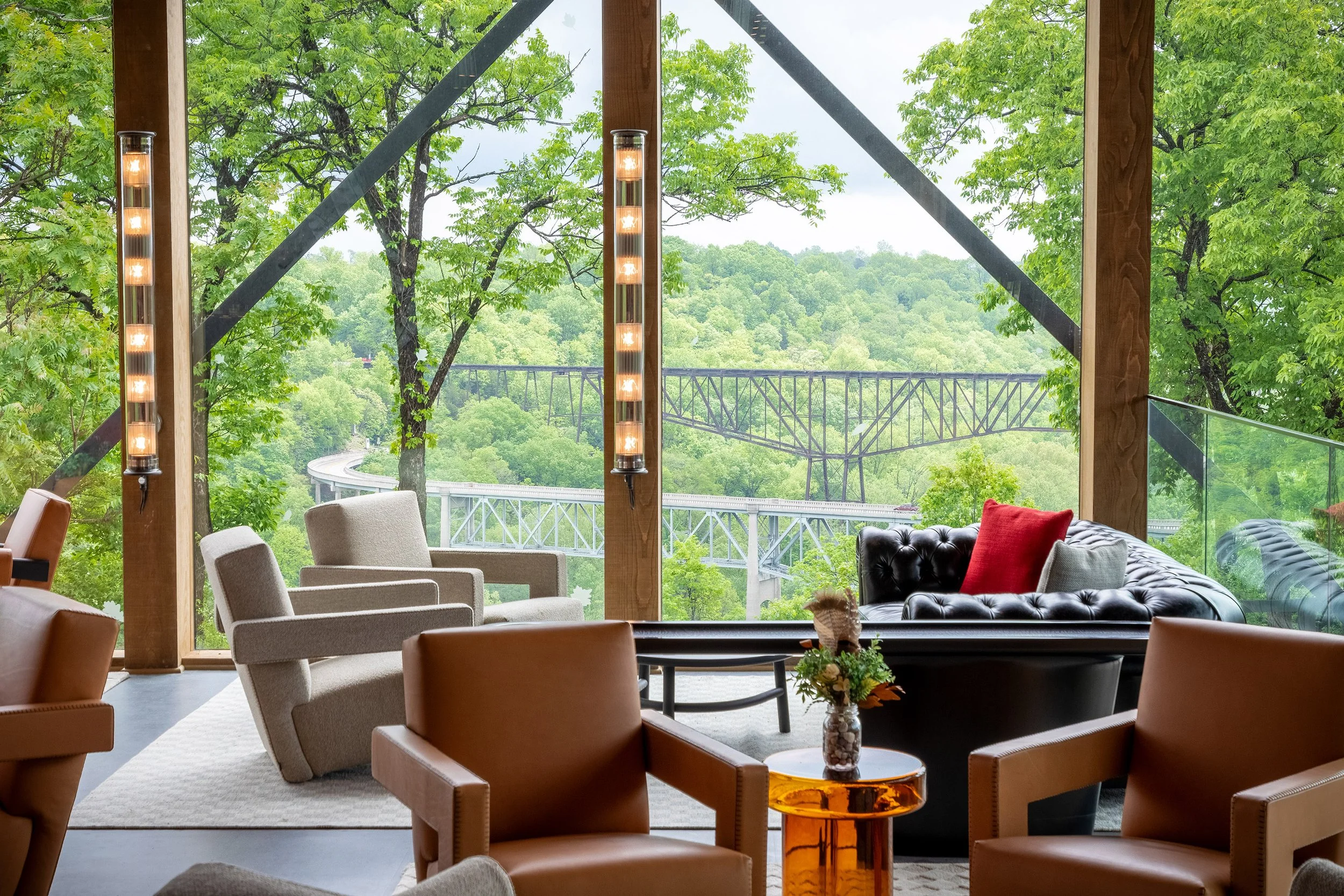 A modern lounge with armchairs, a black tufted sofa, and floor-to-ceiling windows showcases stunning architecture as it overlooks lush green trees and a railway bridge in the distance. Natural light fills the space, creating a cozy and scenic atmosph
