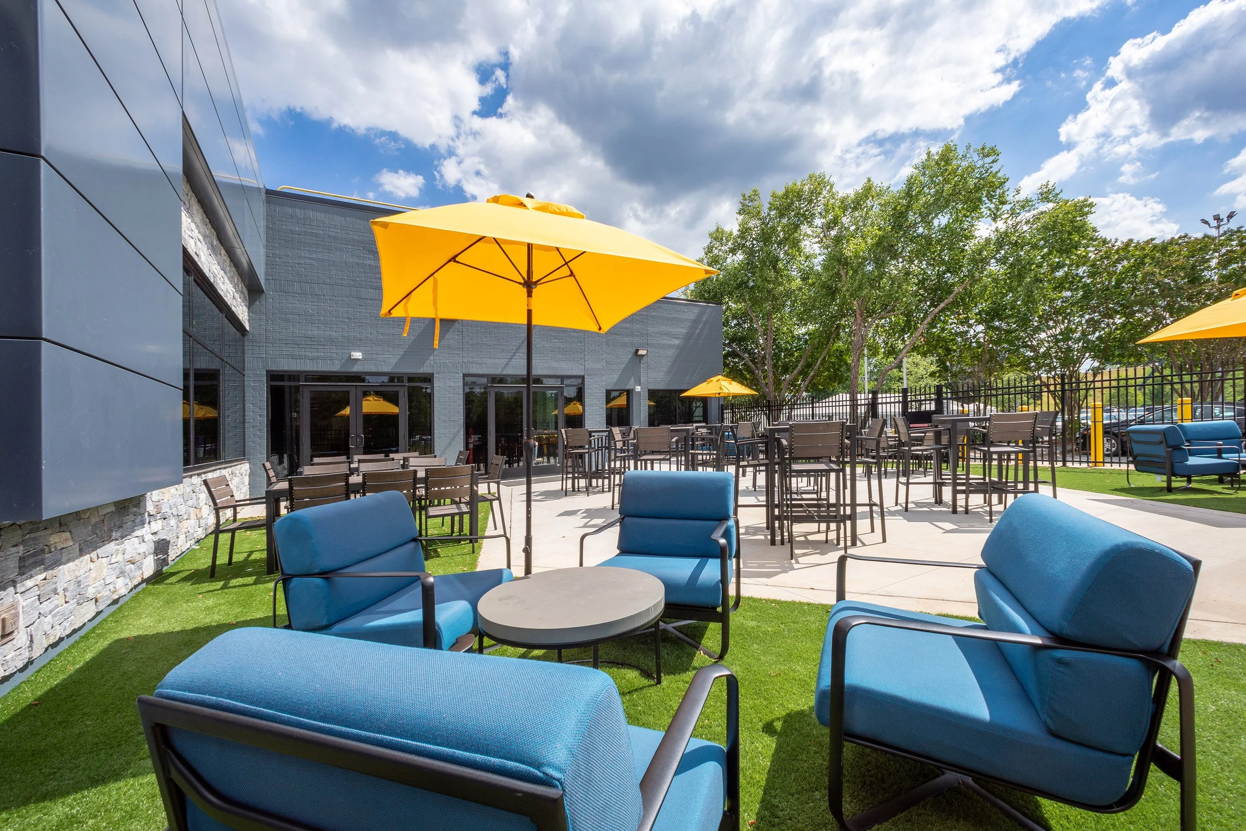 Outdoor patio with blue cushioned chairs and round tables on artificial grass, surrounded by dining tables and yellow umbrellas. Adjacent to a modern building with glass doors.