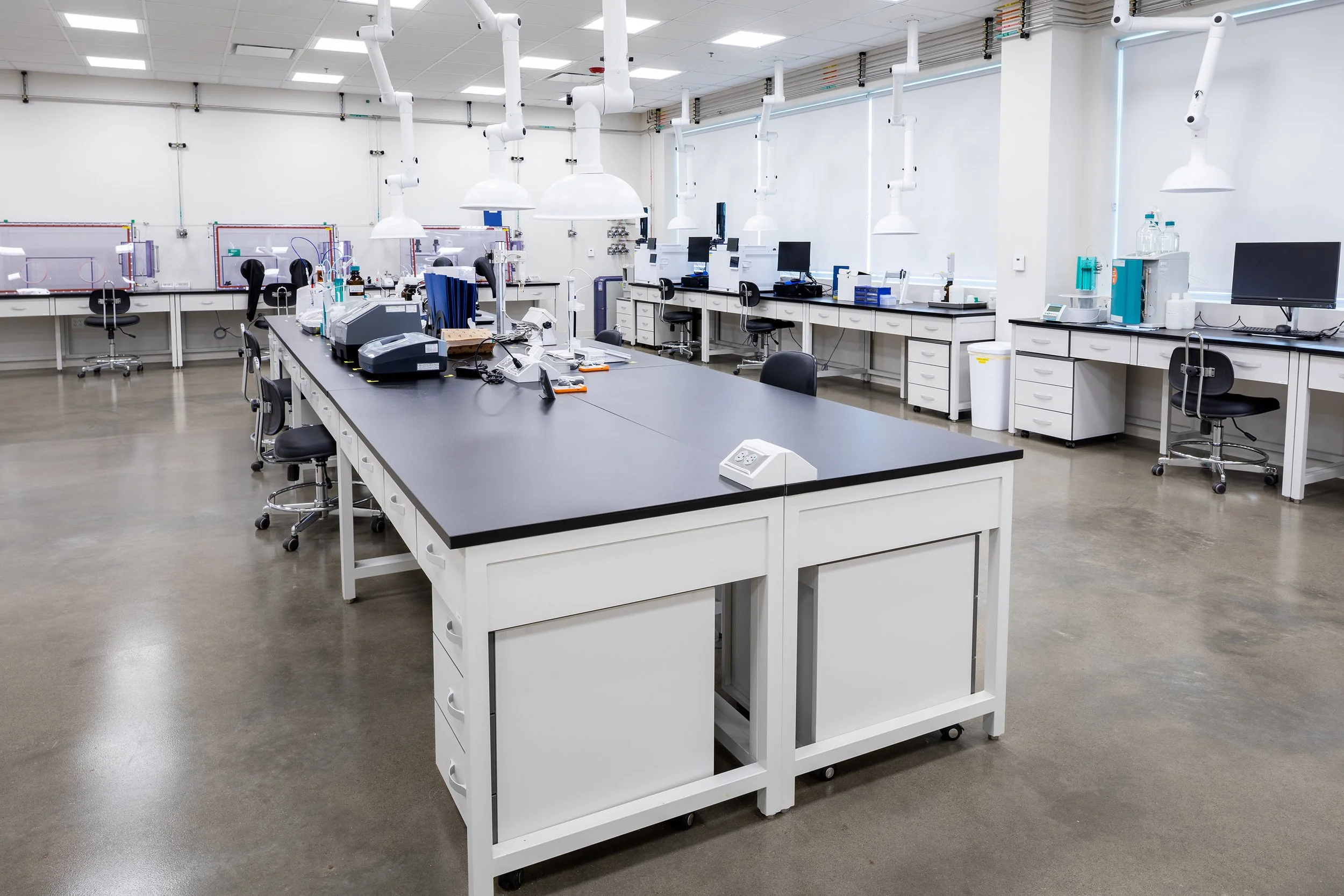 A modern, spacious laboratory with large workbenches, scientific equipment, computers, and lab chairs. The clean, industrial space is well-lit with overhead lights, features white walls, and large windows with blinds.
