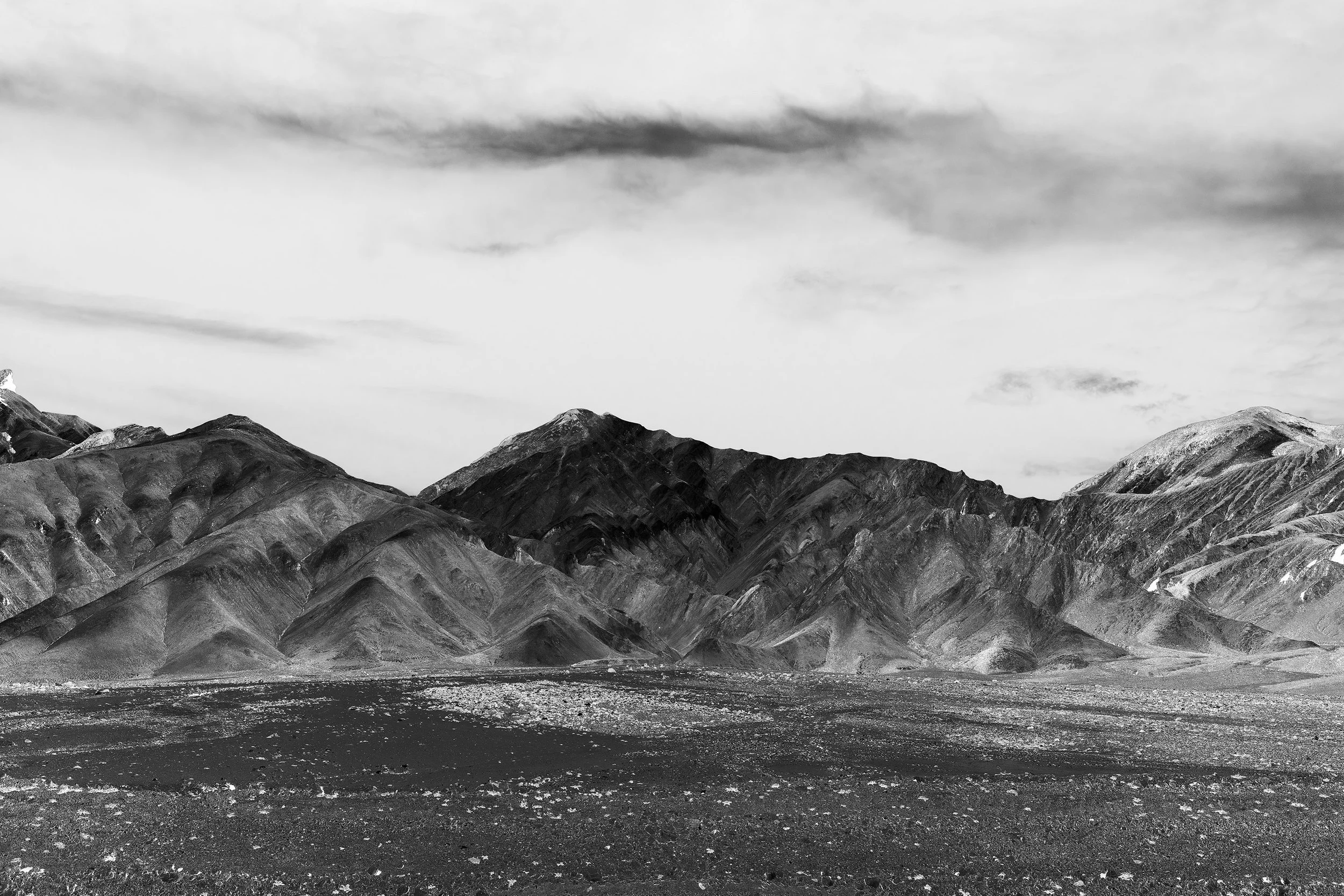 Black and white photo of a mountain range under a cloudy sky, with rocky desert terrain creating an artful landscape in the foreground.
