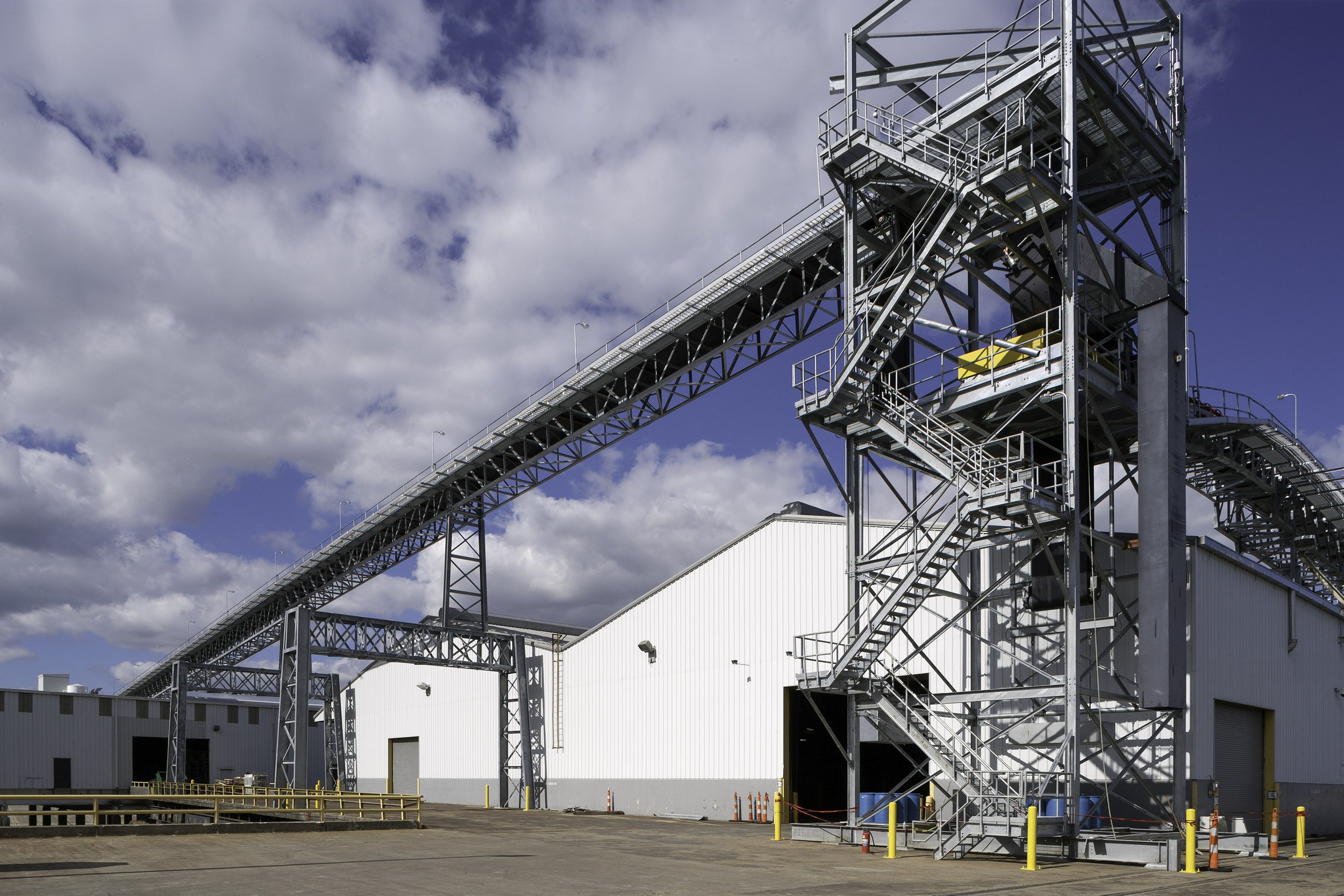 Industrial facility with metal conveyor structure and stairs beside large white warehouse buildings under a partly cloudy sky.