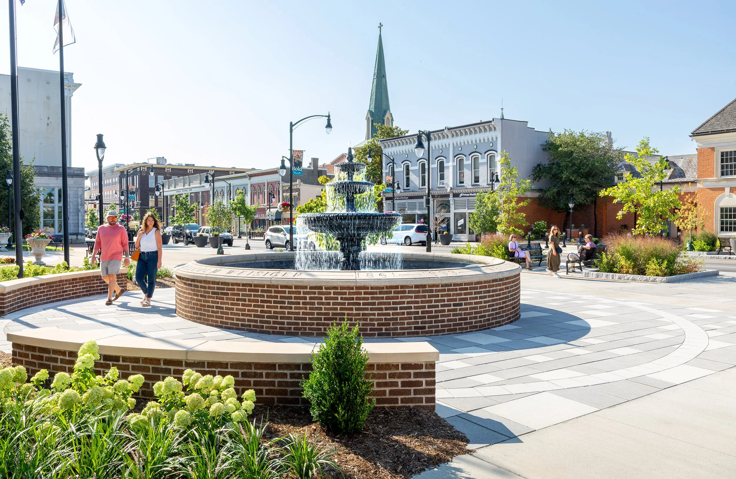 A round brick fountain on the historic main street, surrounded by people and architectural buildings on a sunny day.