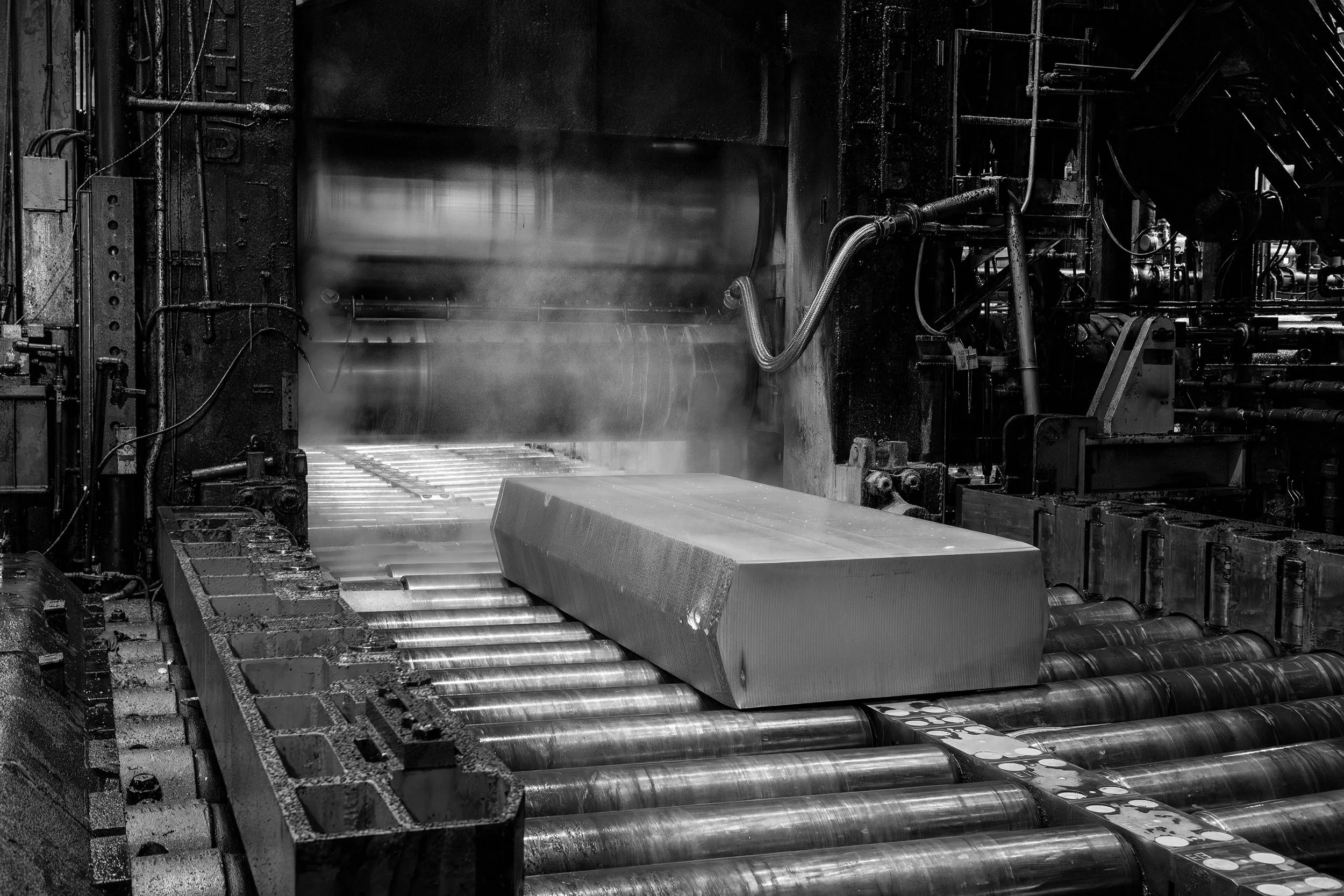 Large aluminum slab on rollers in an industrial factory with steam rising, captured in black and white.