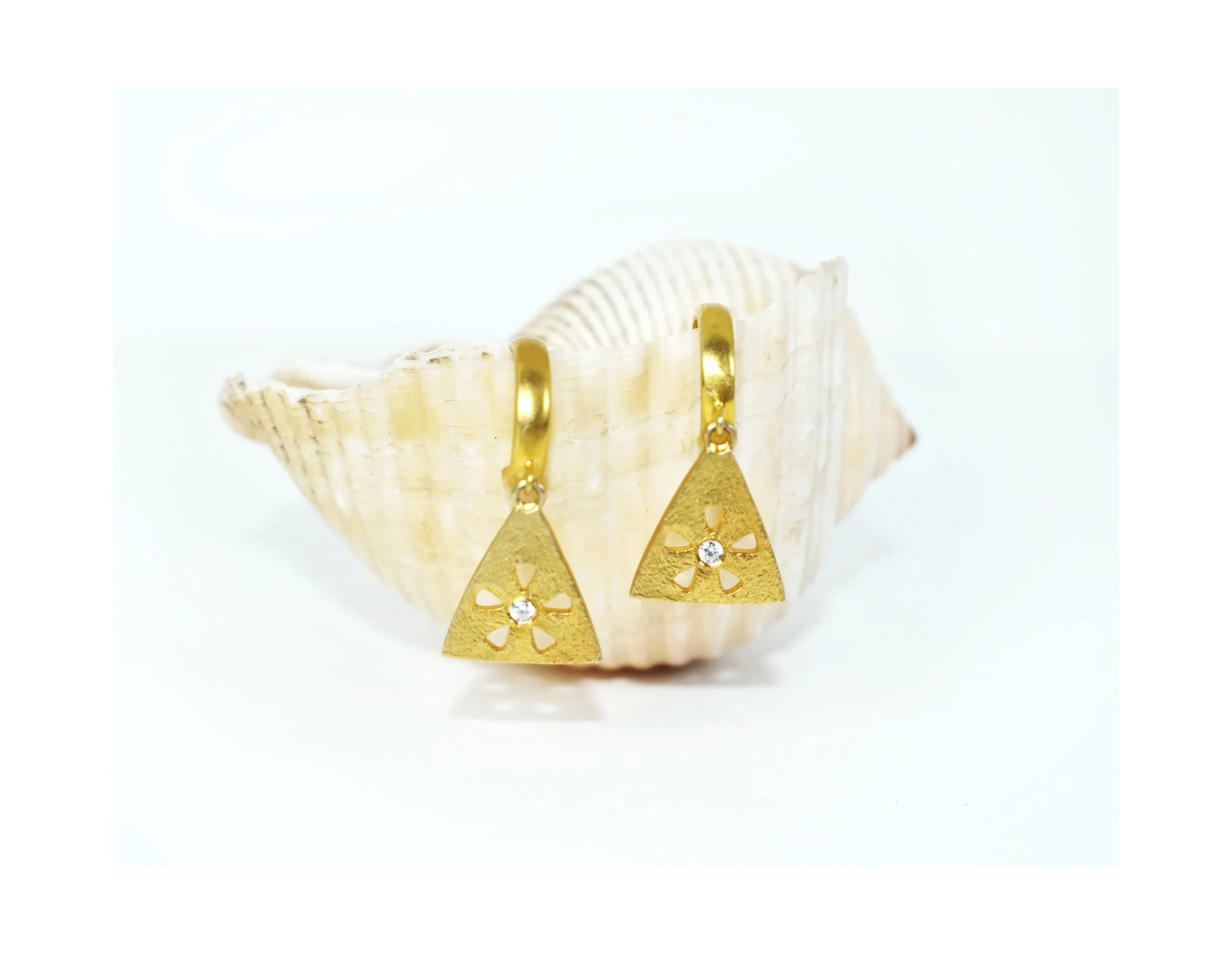 Gold earrings with triangle pendant design and small embedded gemstone, resting on a shell.