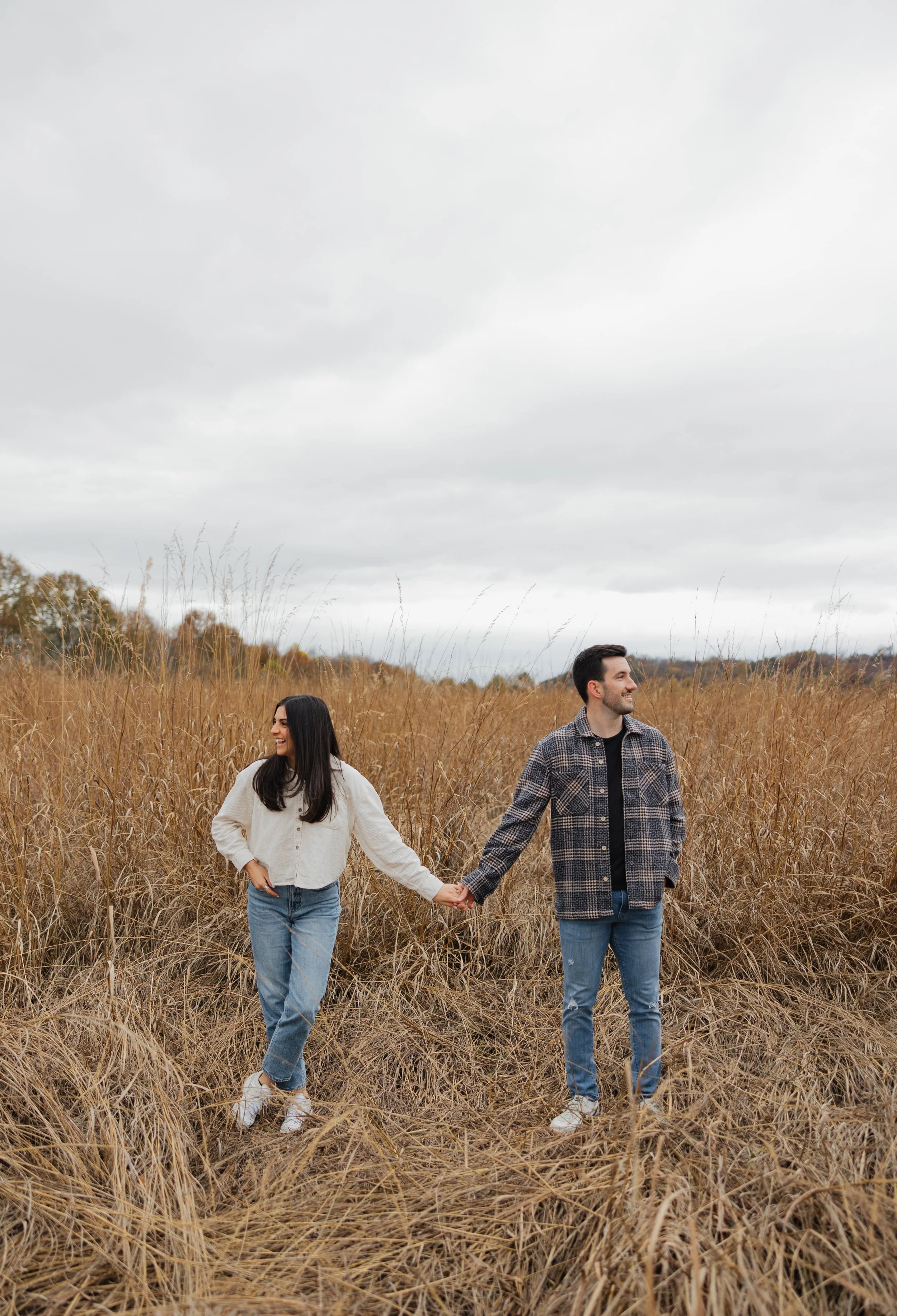 A man and a woman are holding hands and walking through a field of tall, dry grass on a cloudy day, smiling and looking at each other.