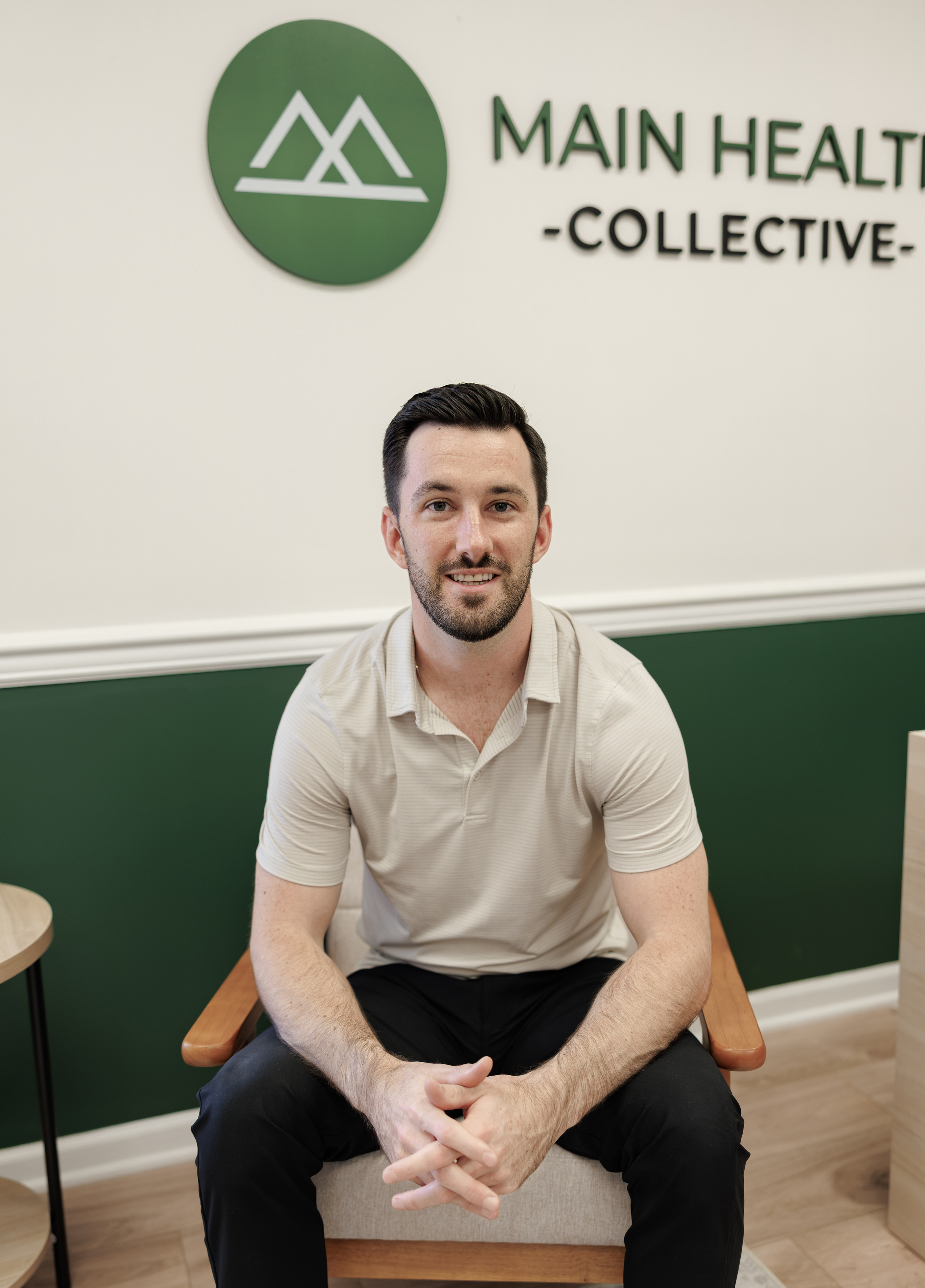 A man with dark hair and a beard, wearing a light beige shirt, sitting on a wooden chair with arms crossed, smiling at the camera. Behind him is a wall with a green and white logo and the text "MAIN HEALTH COLLECTIVE".