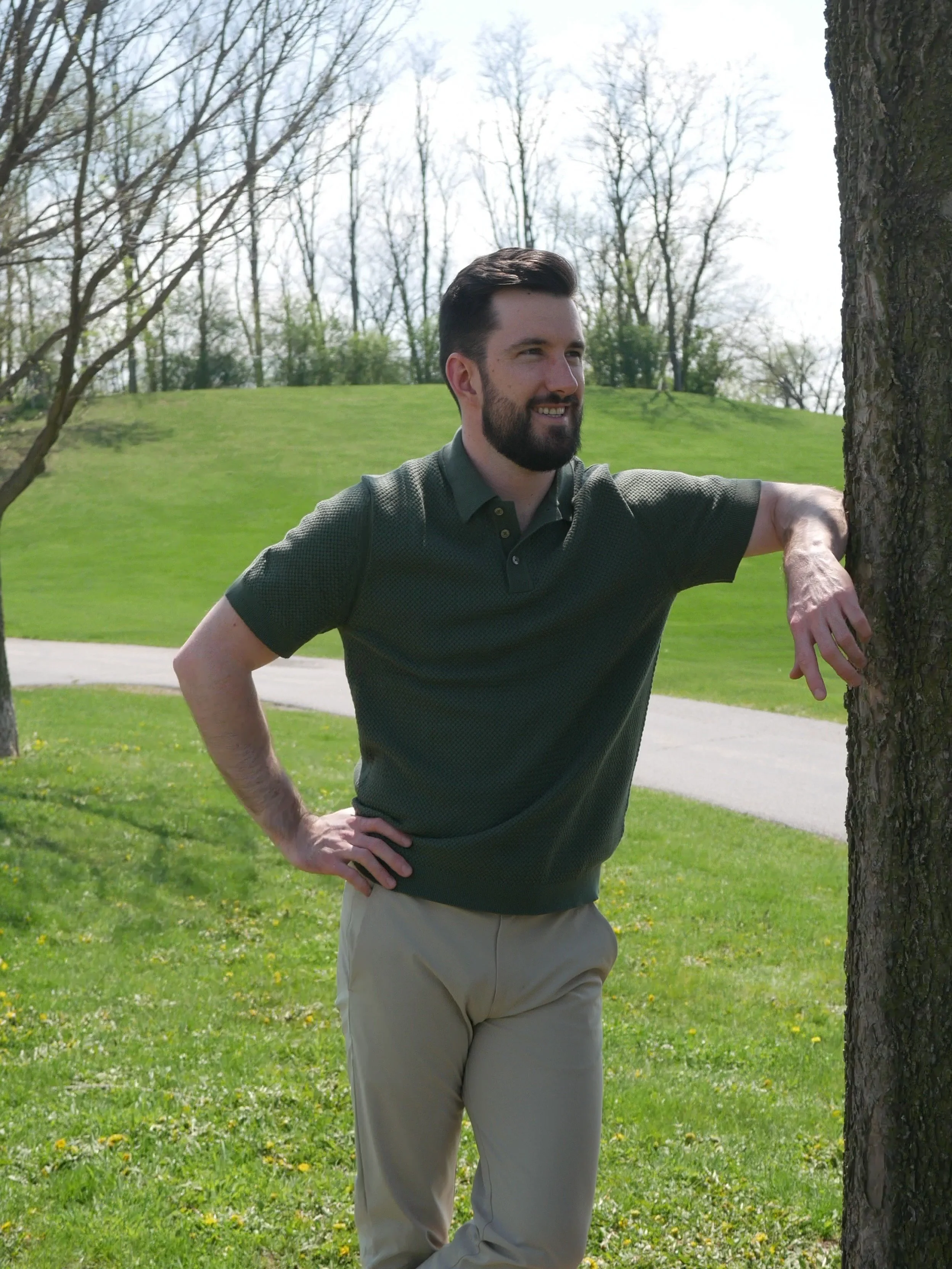 A man with dark hair and a beard smiling while leaning against a tree outdoors in a park with green grass and leafless trees in the background.