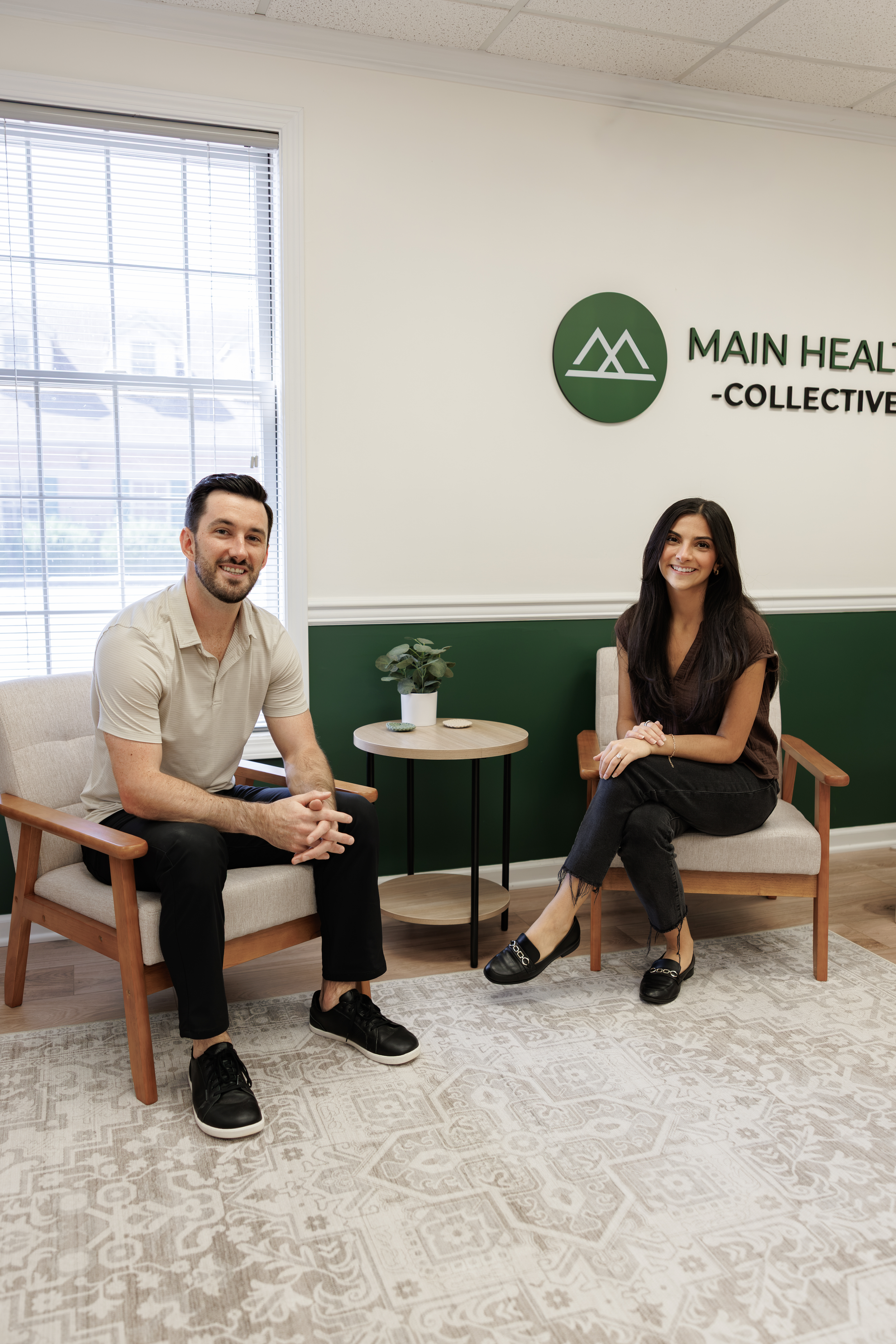 A man and woman sitting on chairs in an office with a sign reading "Main Health Collective" on the wall behind them. There is a window with blinds, a small round table with a plant, and a decorative rug on the floor.