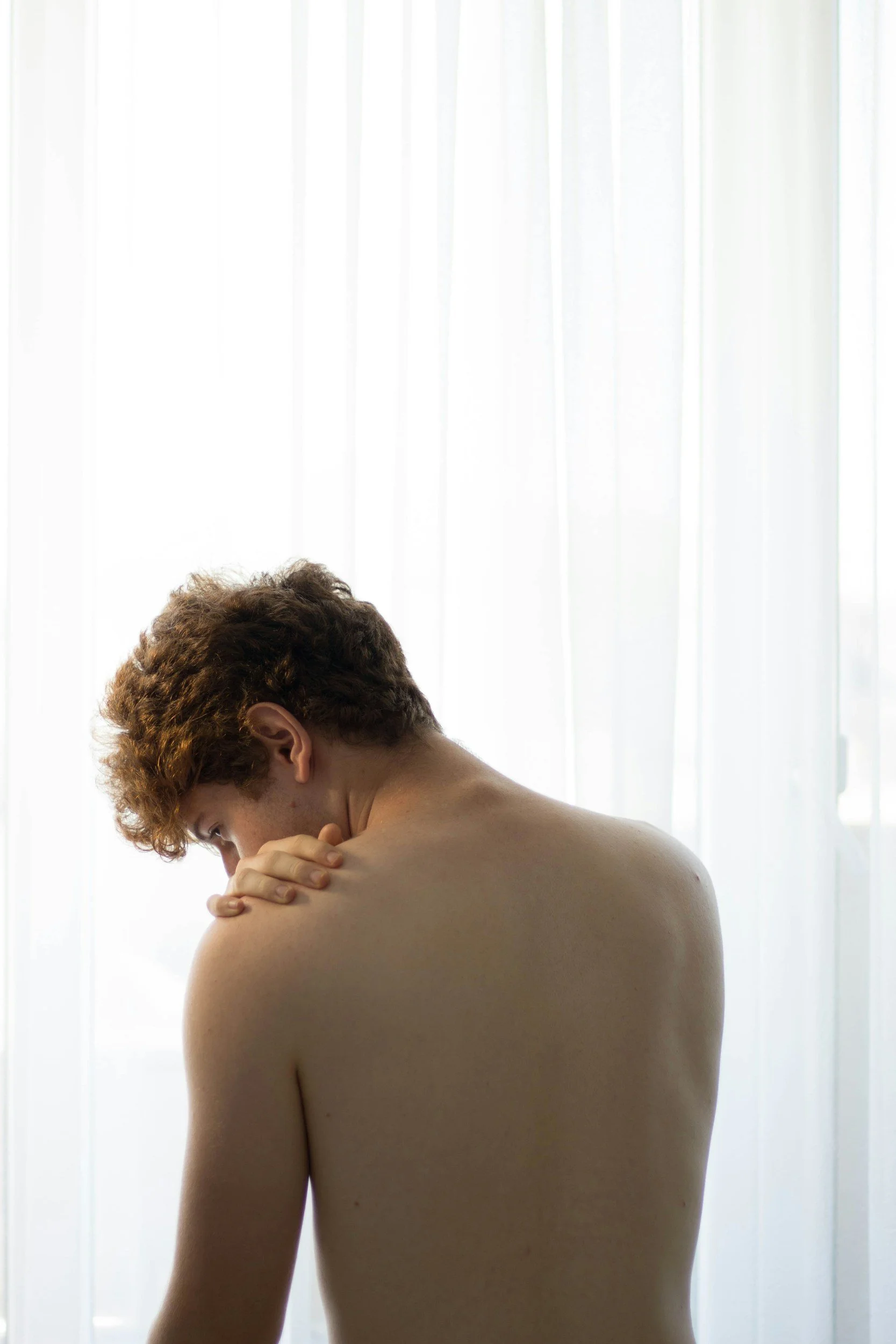 A young man with curly red hair, shirtless, standing in front of a white curtain, with his head bowed and hand resting on his shoulder.