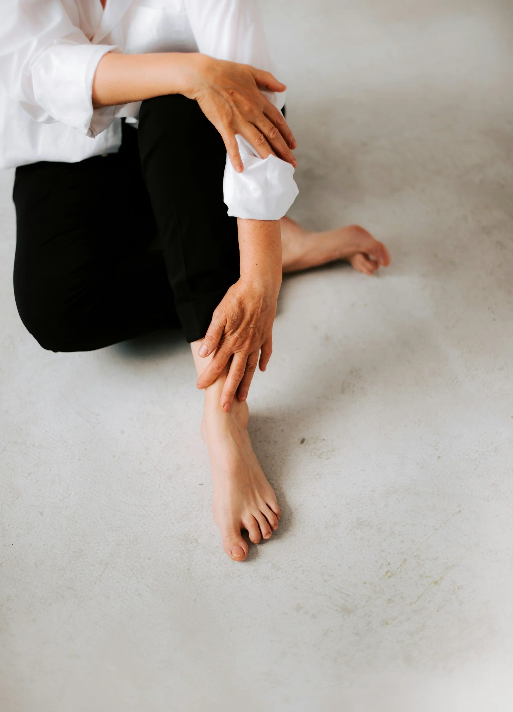 A person sitting on the floor, wearing a white shirt and black pants. One hand is resting on their knee, and a second hand is holding and supporting a person's foot by the ankle, with the foot extended outward. The background is a plain, light-colored surface.