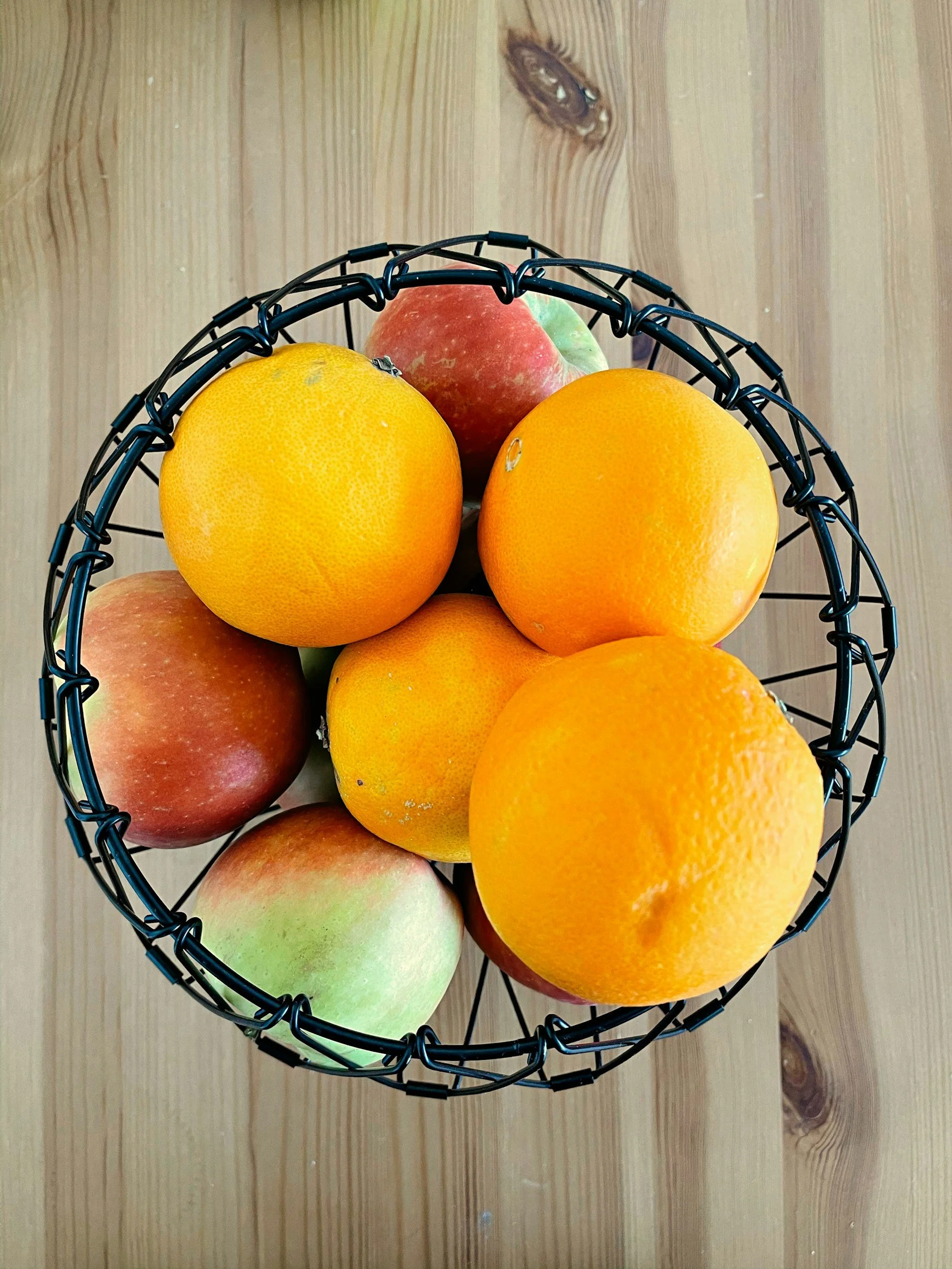 A top view of a black wire basket filled with oranges and apples on a wooden surface.