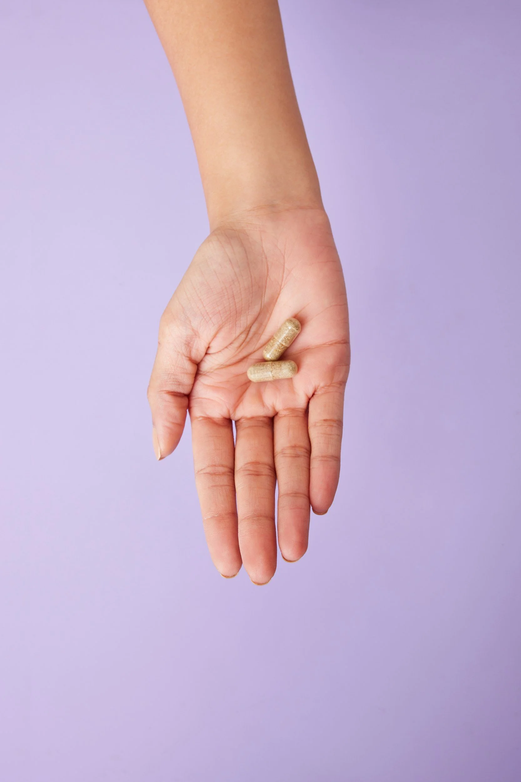 Two hands holding three capsules against a purple background.