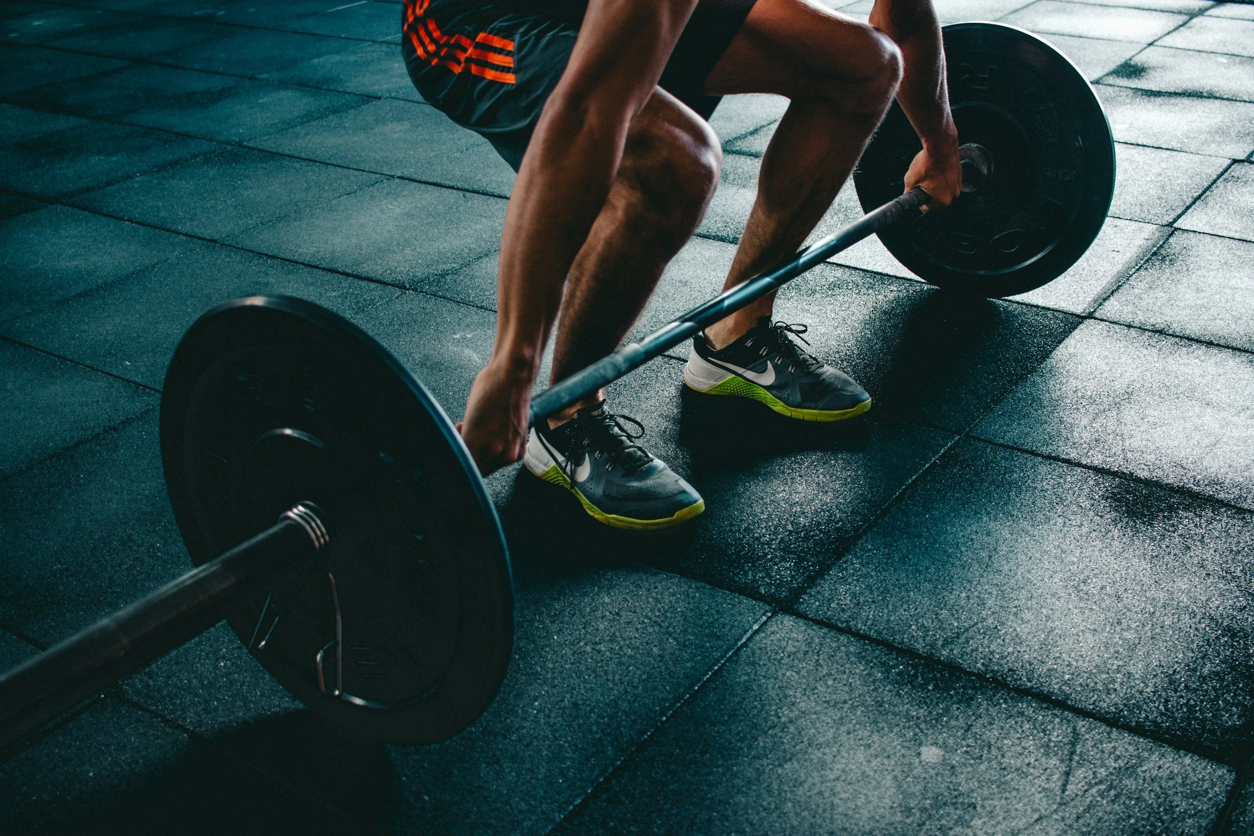 A person in athletic shoes and shorts is lifting a barbell on a gym floor.