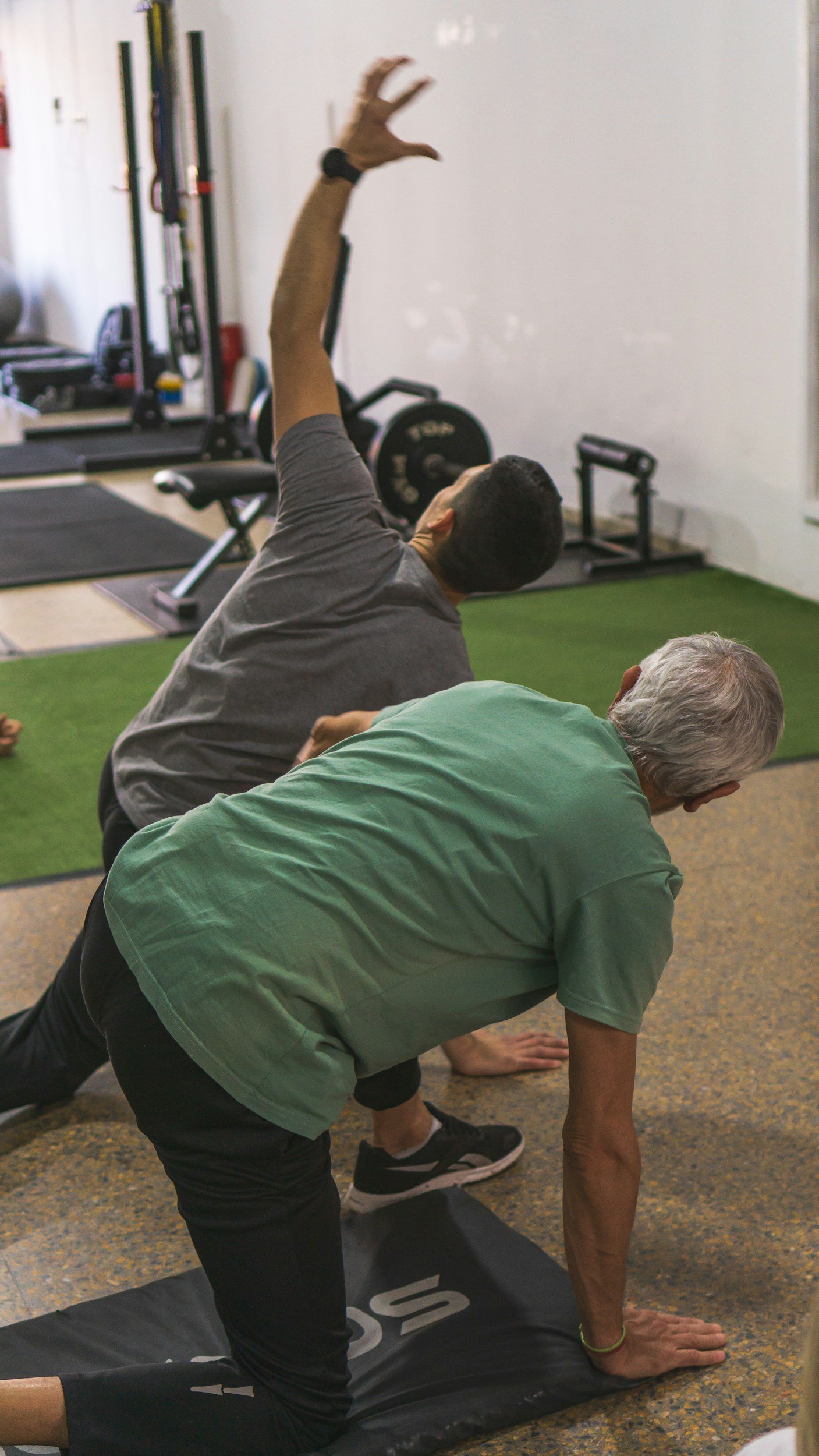 Two people in workout attire practicing yoga or stretching in a gym, with exercise equipment visible in the background.