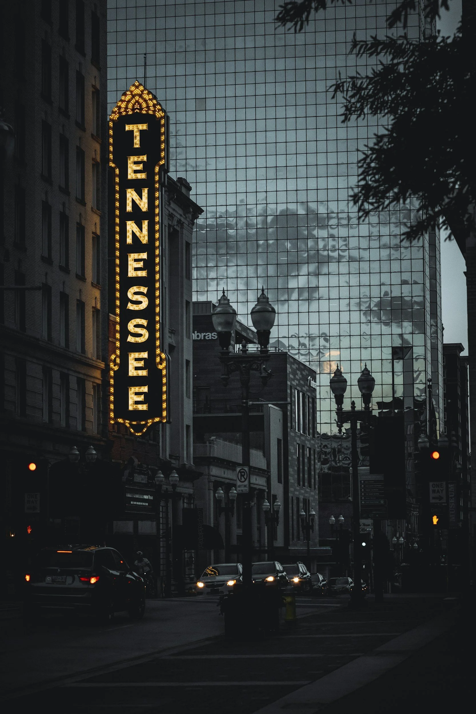 City street scene during dusk with illuminated sign reading 'TENN ESEE' on a building, cars on the street, and a reflective glass skyscraper in the background, partially obscured by tree branches.