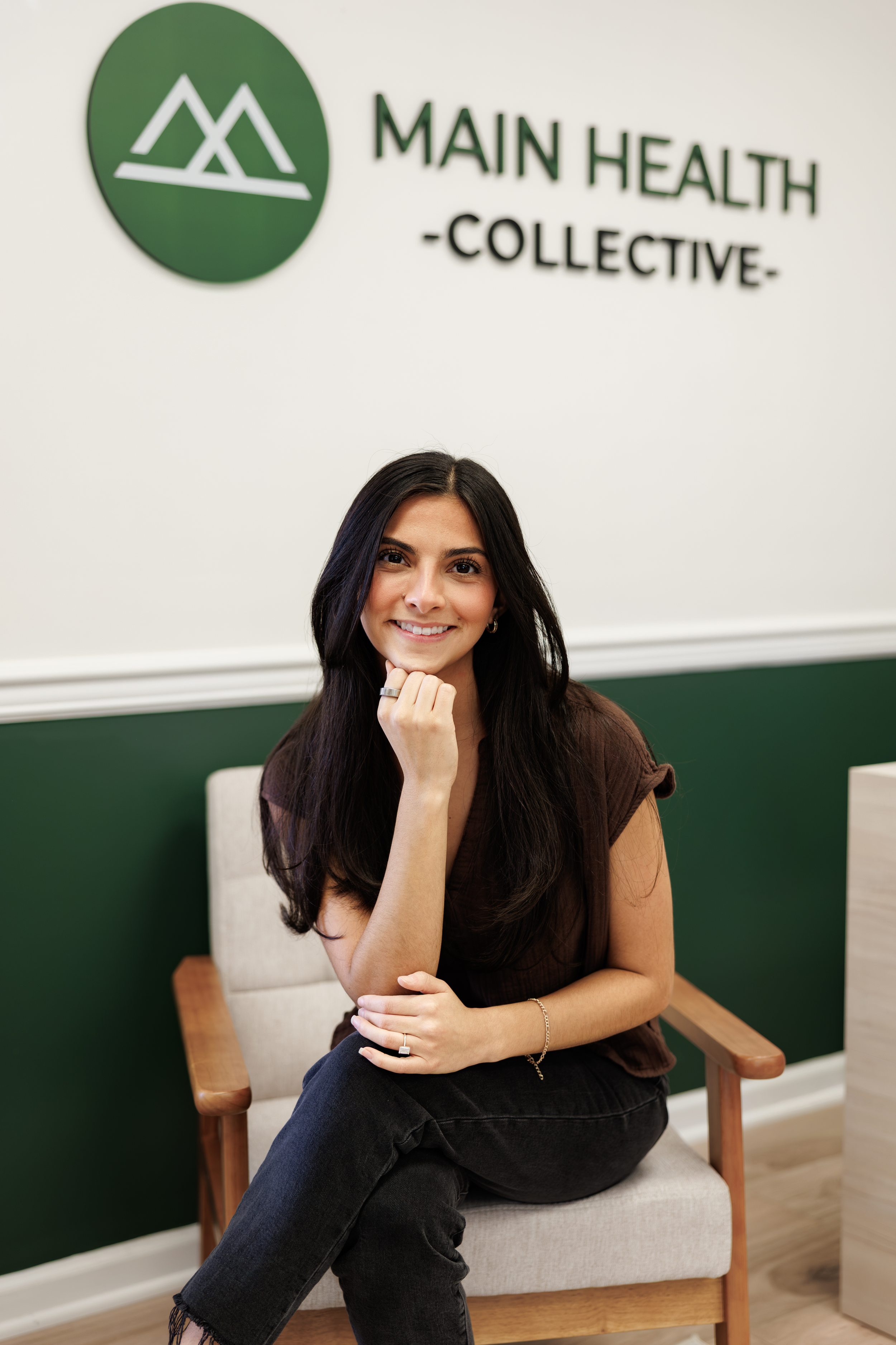 A woman sitting in a wooden armchair with beige cushion, smiling, with a green and white sign on the wall behind her that reads "Main Health Collective".