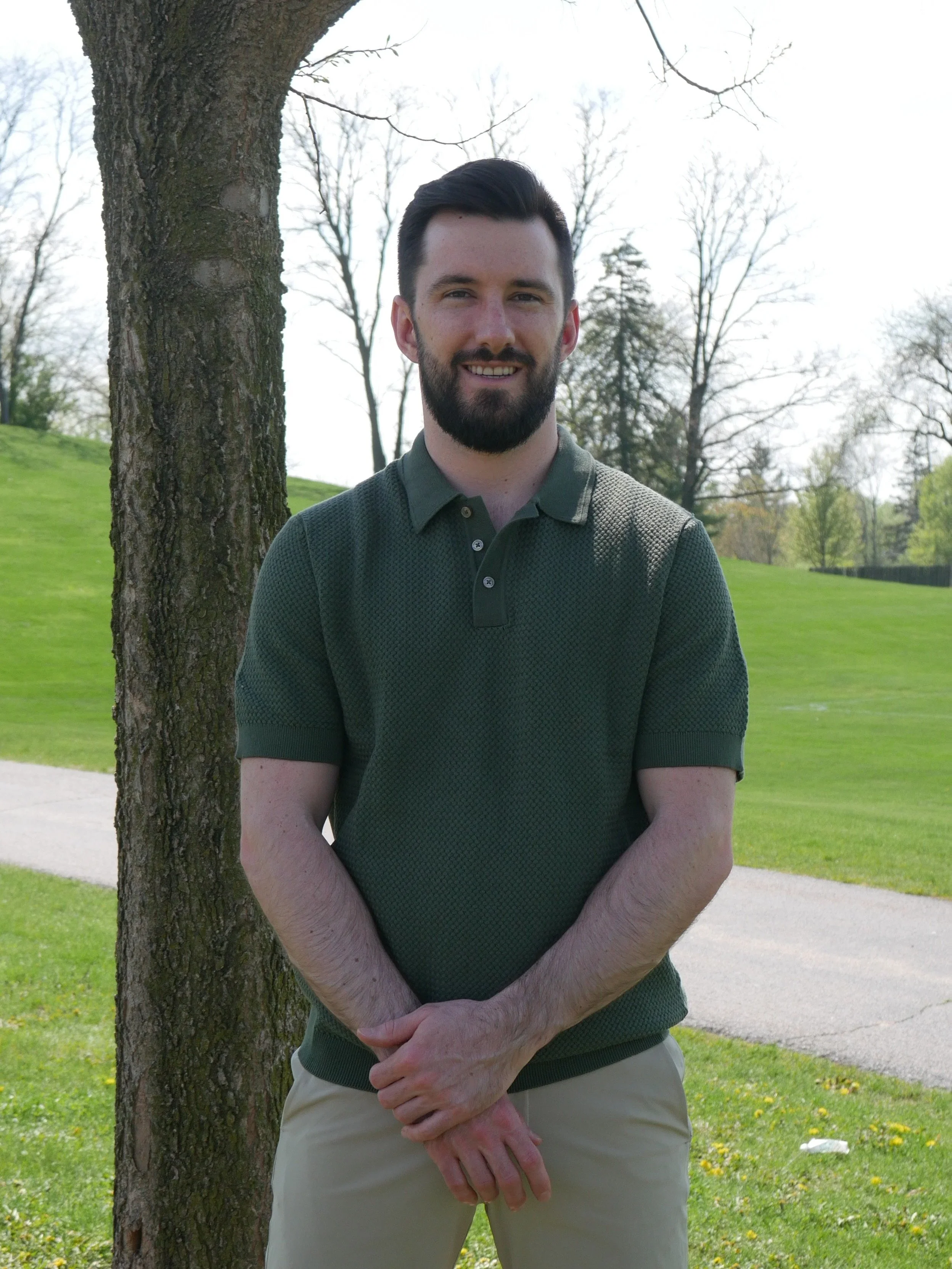 A man with dark hair and a beard standing next to a tree in a park with green grass and leafless trees in the background on a clear day.