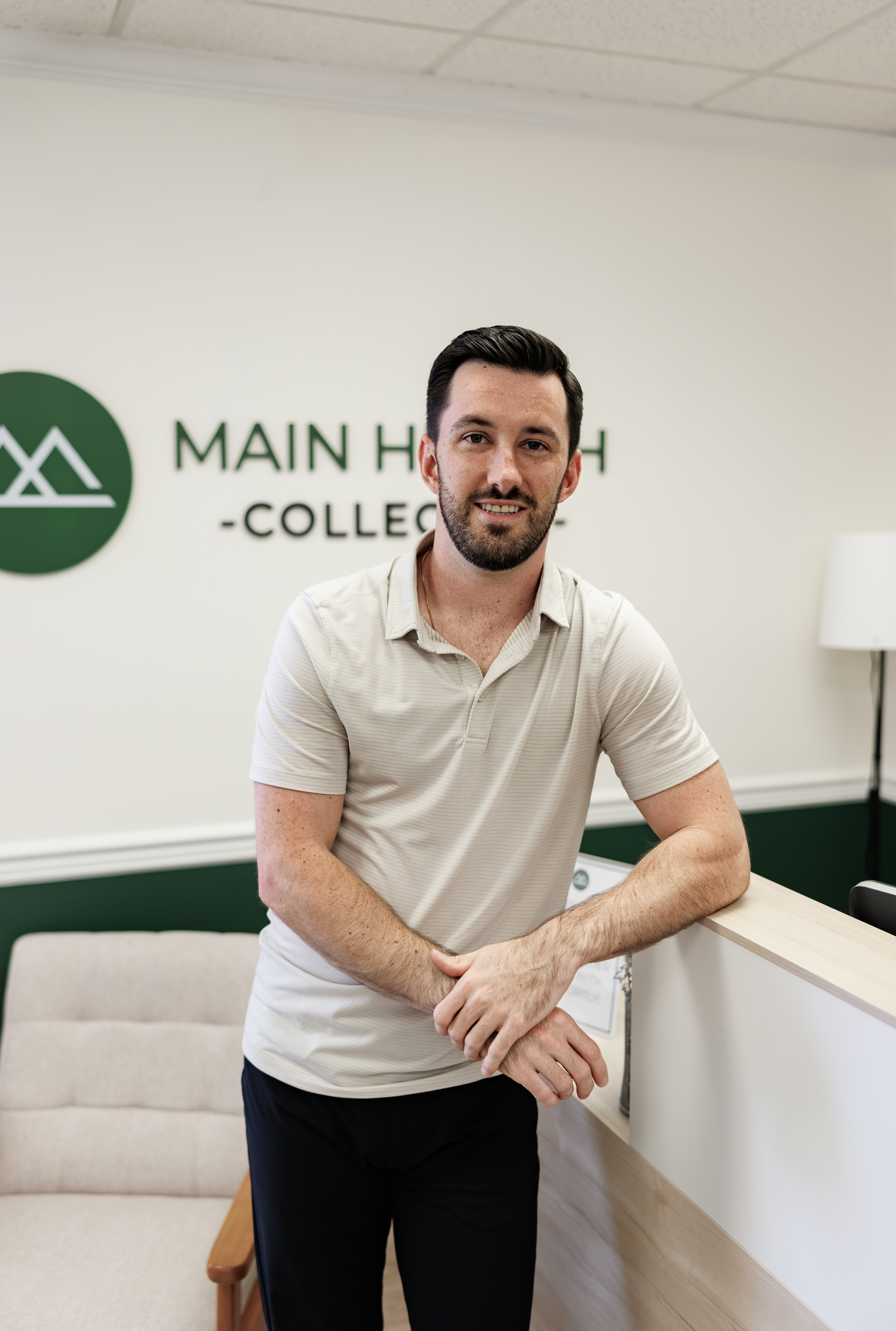 A young man with dark hair, a beard, wearing a light-colored striped polo shirt, leaning on a reception desk, smiling at the camera. Behind him, there is a sign that reads 'Main Health - College' with a green circle logo.