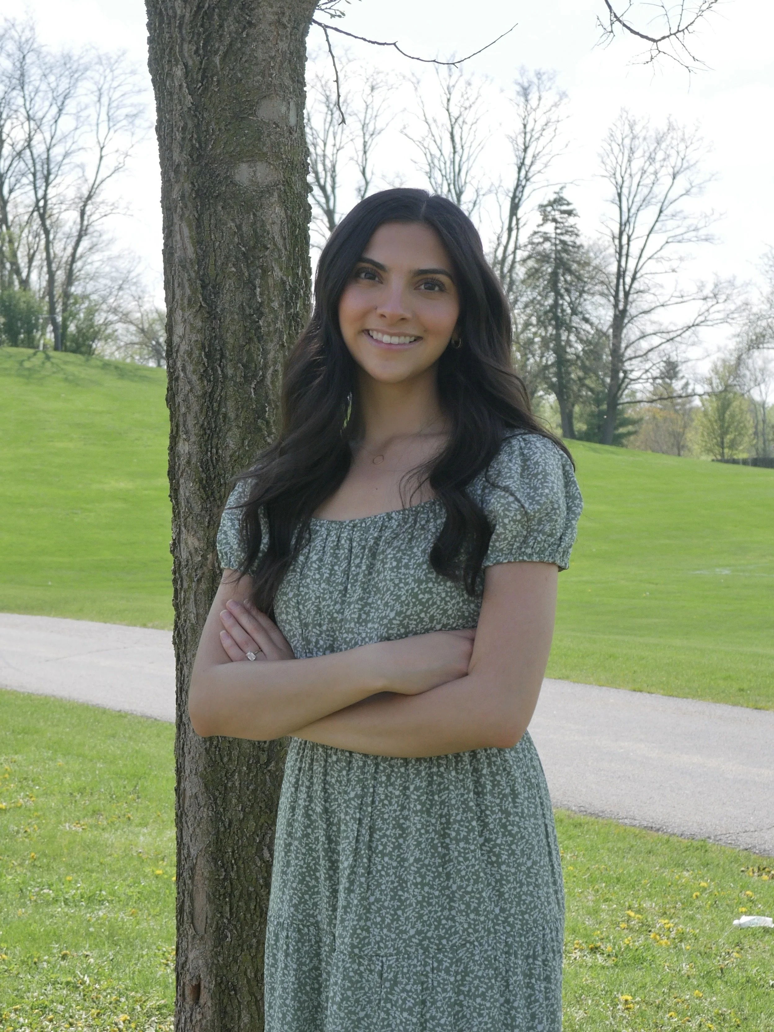 A woman with long dark hair smiling with arms crossed, standing outdoors next to a tree with green grass and trees in the background.