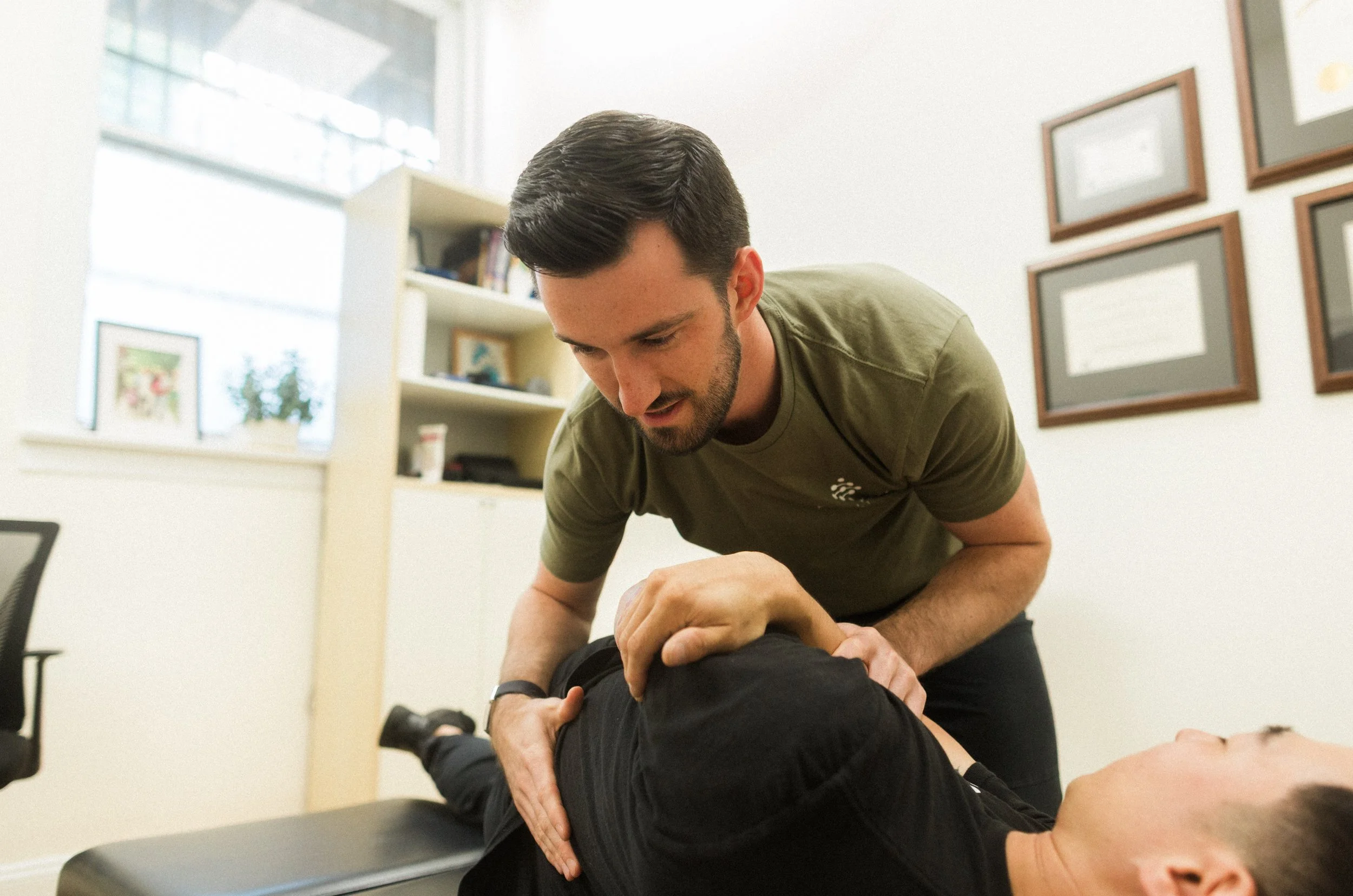A man providing chiropractic adjustments to a patient lying face down on an examination table in a well-lit office with framed pictures on the wall.